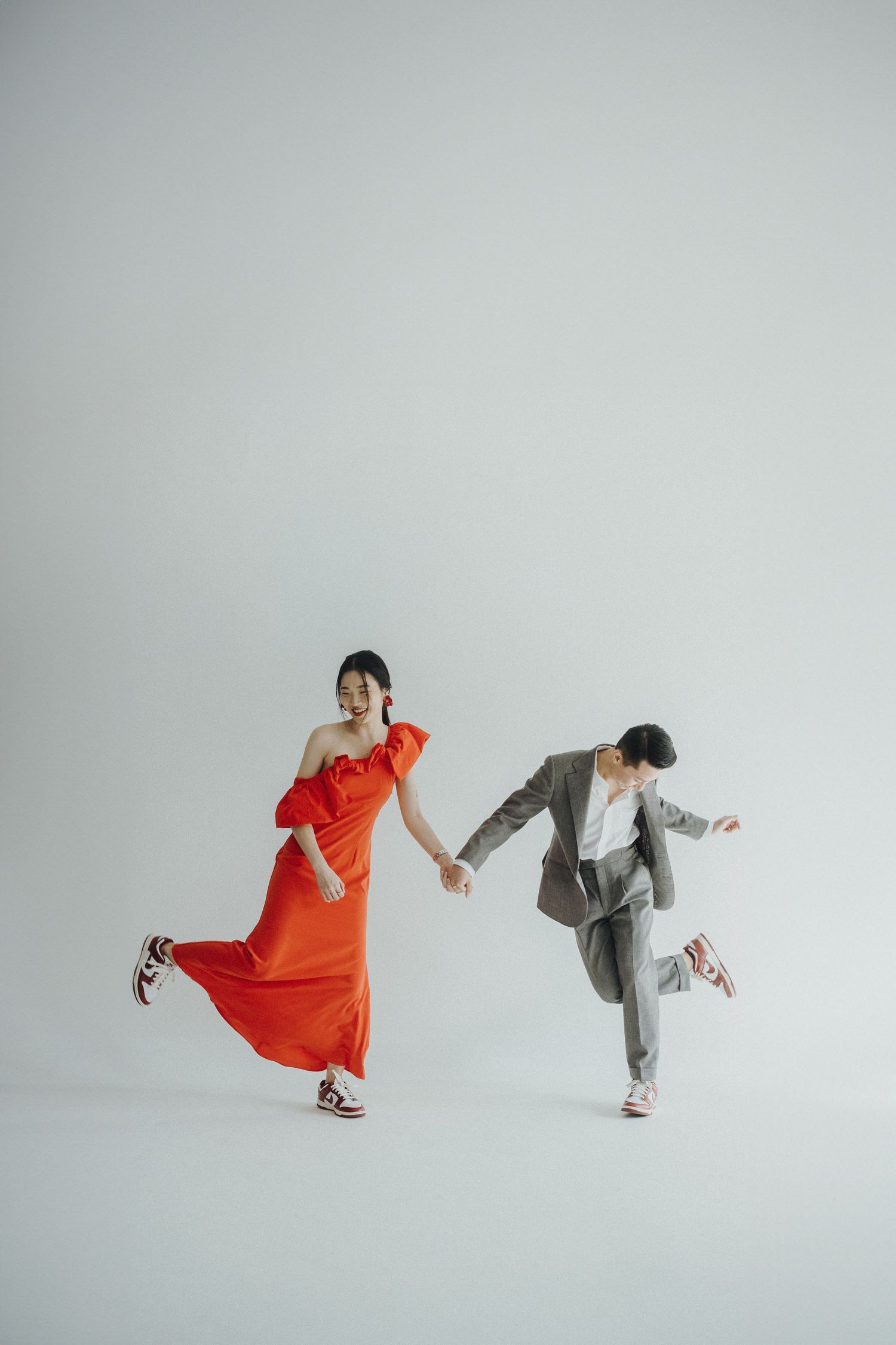 Dancing couple in red dress and gray suit create energetic poses in white studio space.