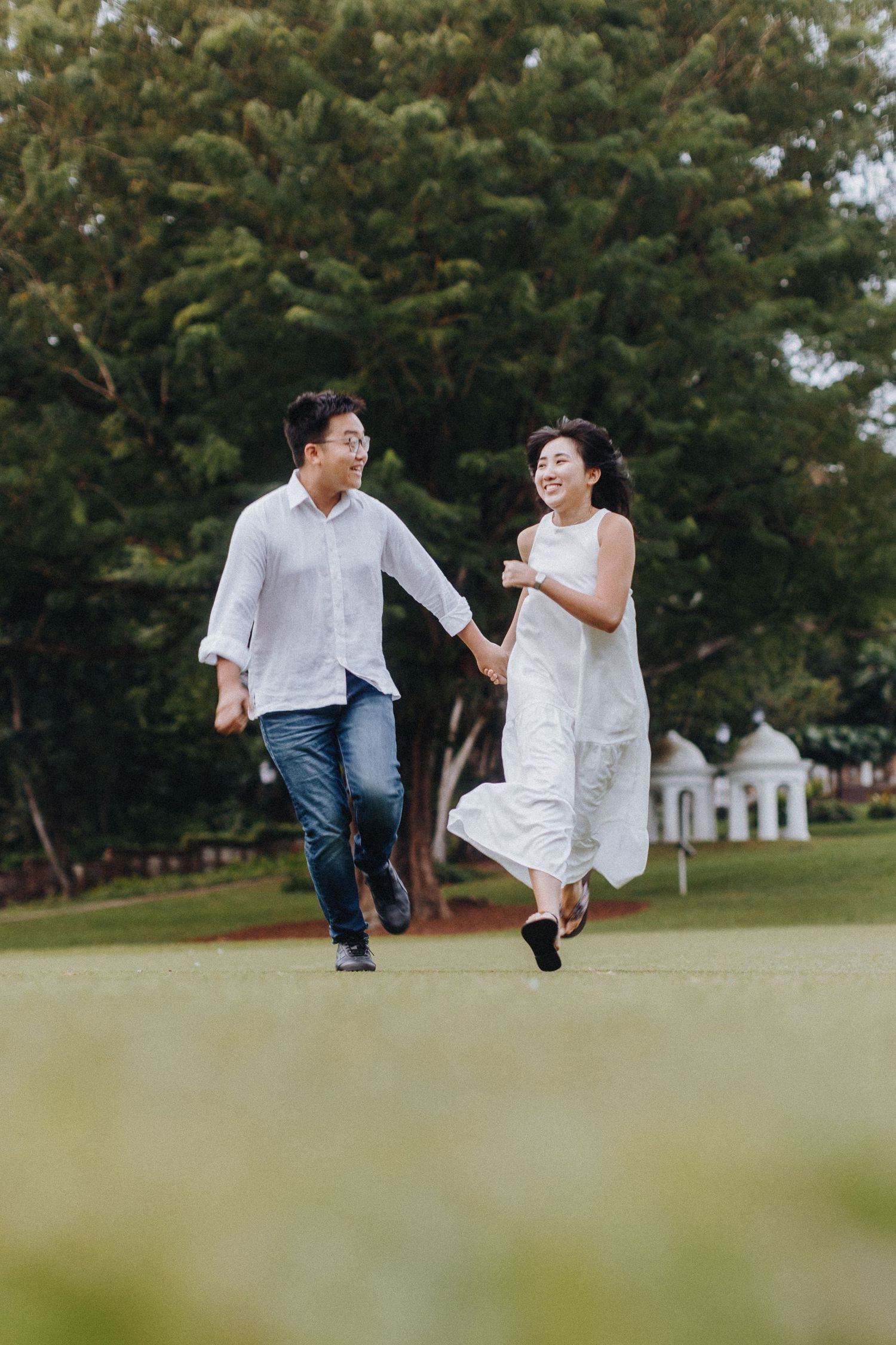 A couple playfully runs and poses in a grassy park area surrounded by trees.