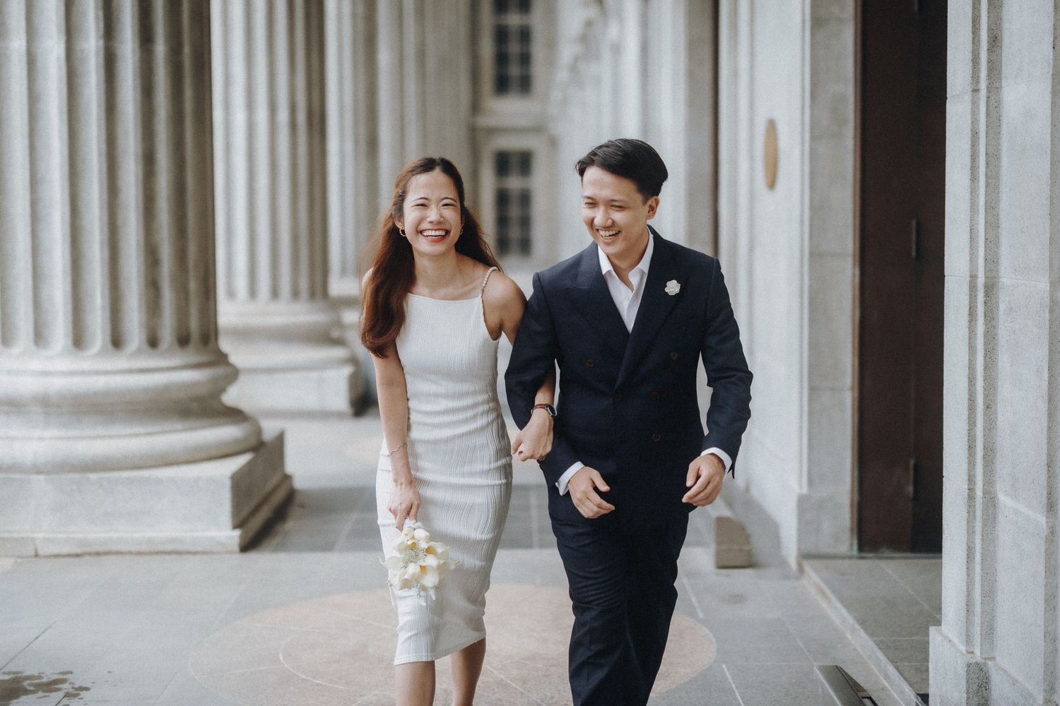 Couple strolls playfully through a classical colonnade with white pillars.