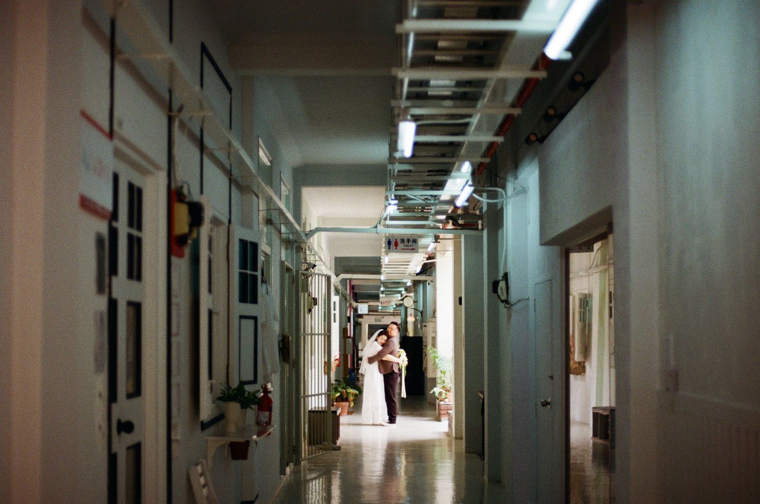 A long corridor with fluorescent lighting and white walls leading to a bright doorway at the end.