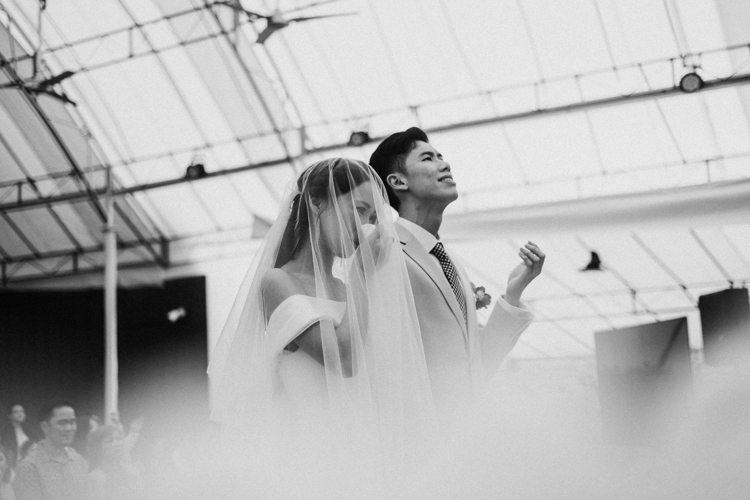 A black and white candid wedding photo captured under an industrial ceiling with dramatic lighting.