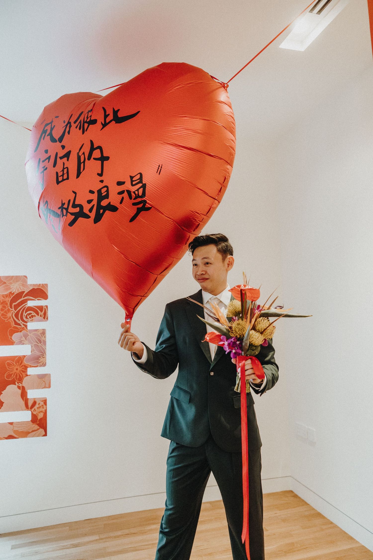 Groom in dark suit holding large red heart balloon and flowers during wedding celebrations.