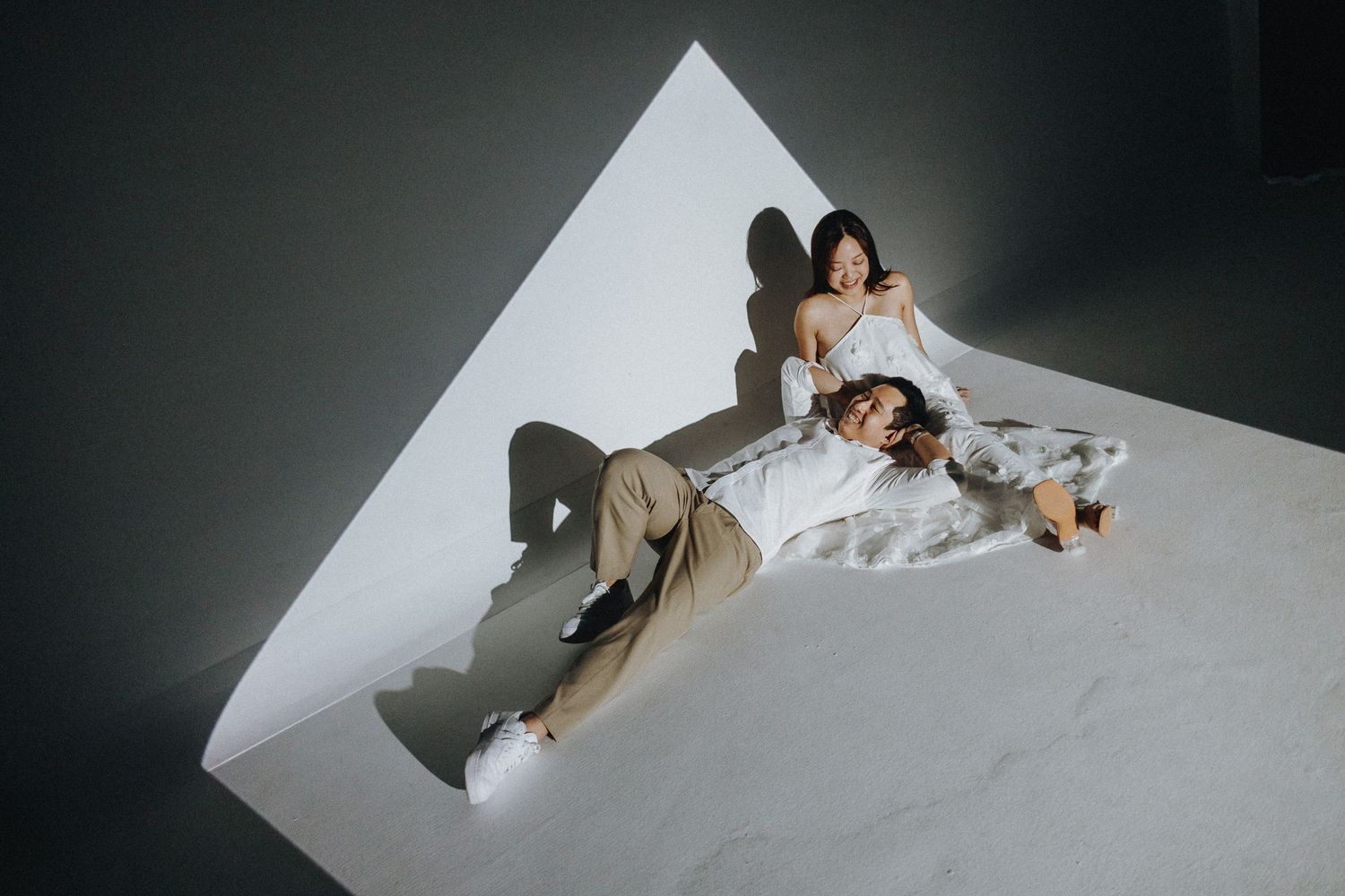 Two people pose artistically on angled white surface with dramatic lighting and shadows creating geometric shapes.