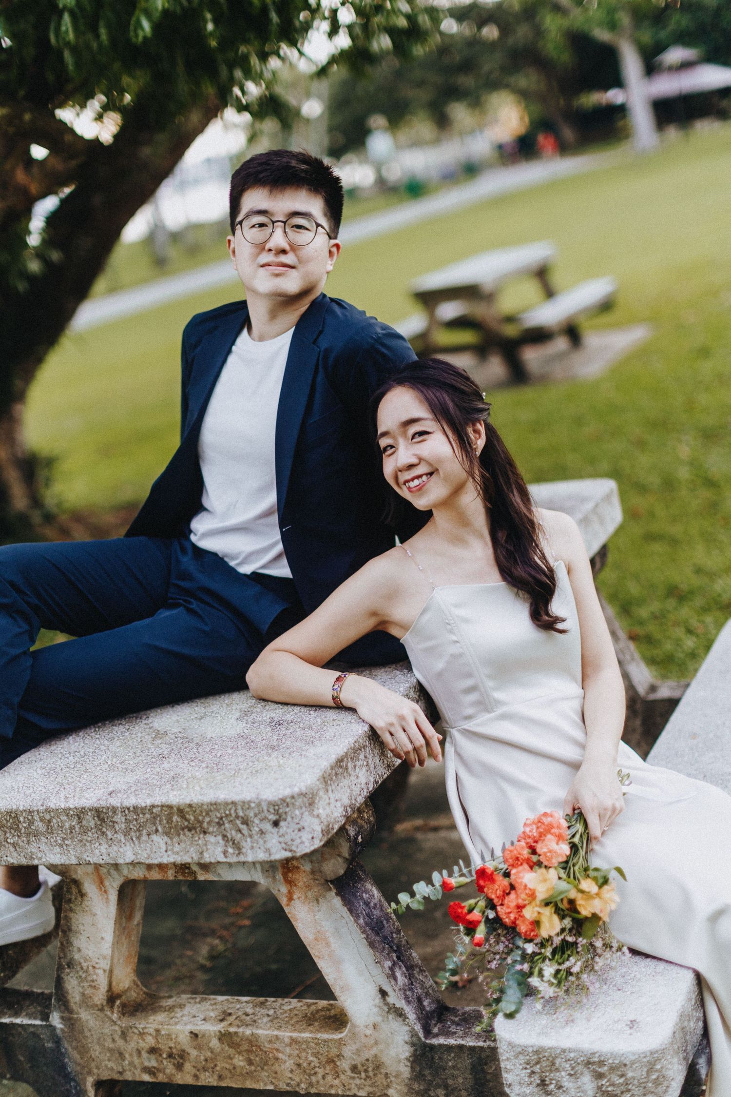 A couple sits together on a stone picnic bench in a park, the bride holding a coral and peach bouquet.