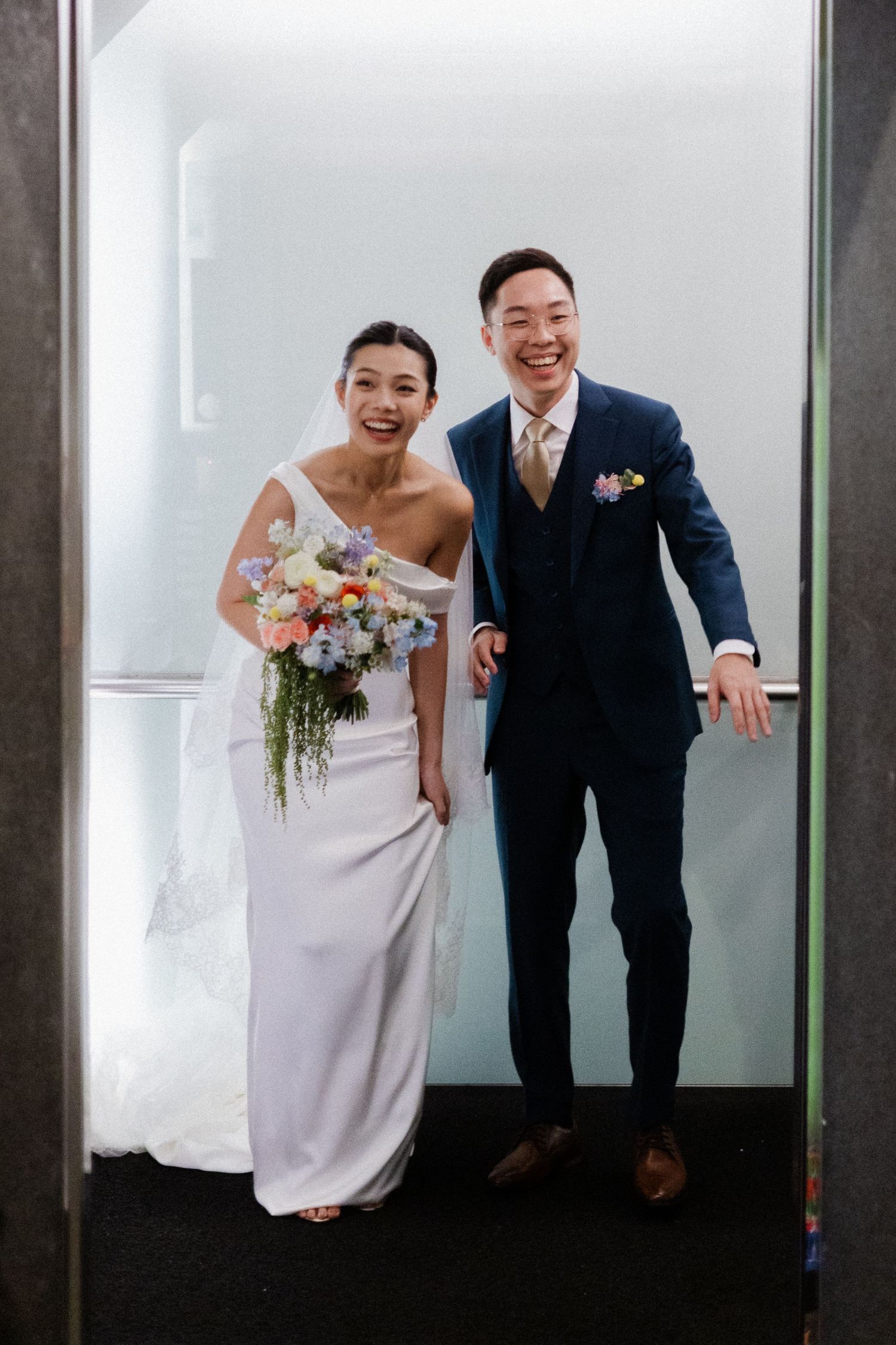 Happy bride and groom posing together in elevator, bride holding colorful bouquet, groom in navy suit.