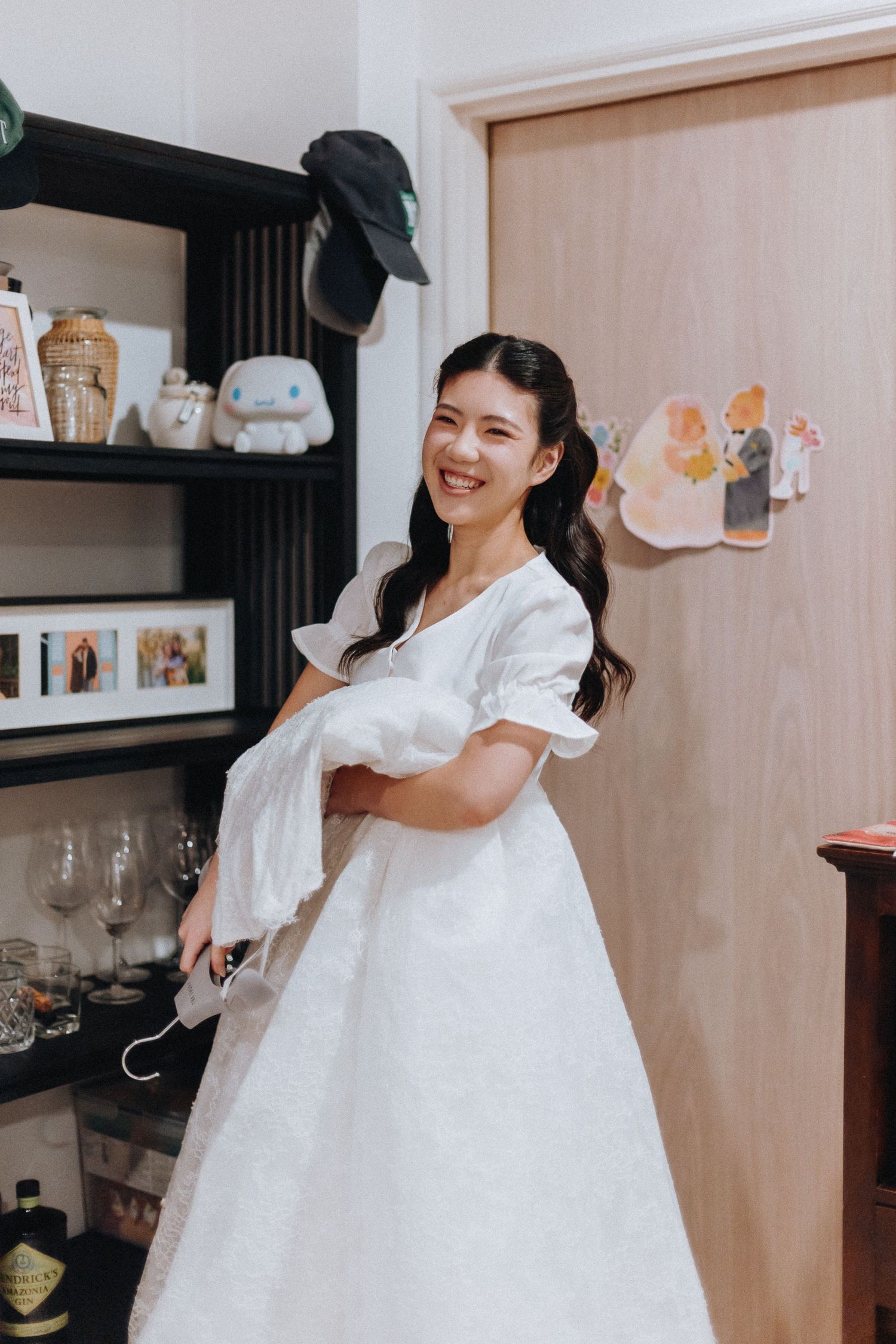 Smiling Asian woman in white wedding dress holding veil, standing near bookshelf with decor in cozy room.
