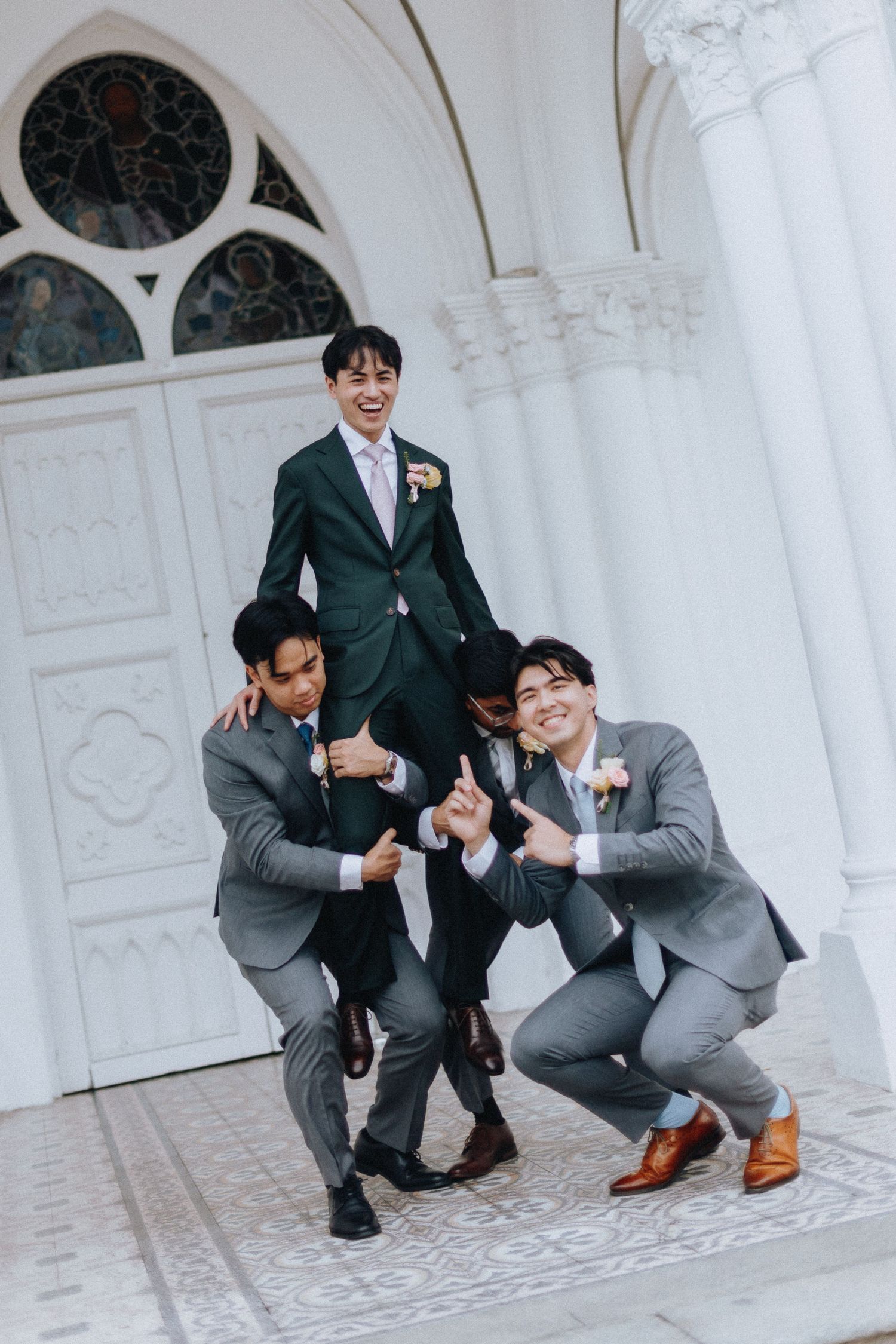 Groom in dark suit lifted by two groomsmen in grey suits posing playfully outside white church doors.