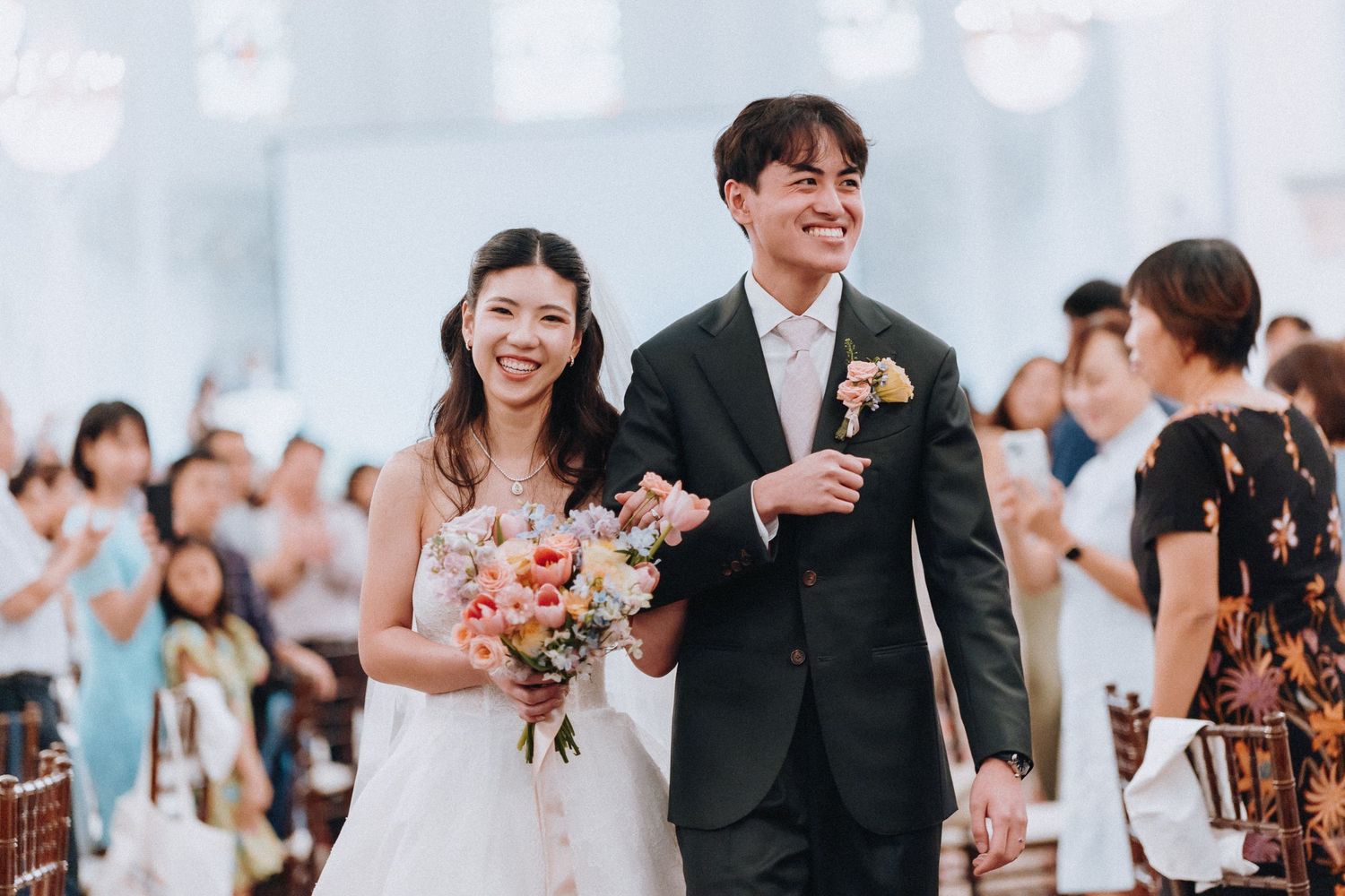 Happy newlyweds walk down the aisle smiling, bride holding colorful bouquet, groom in dark suit, guests applauding.