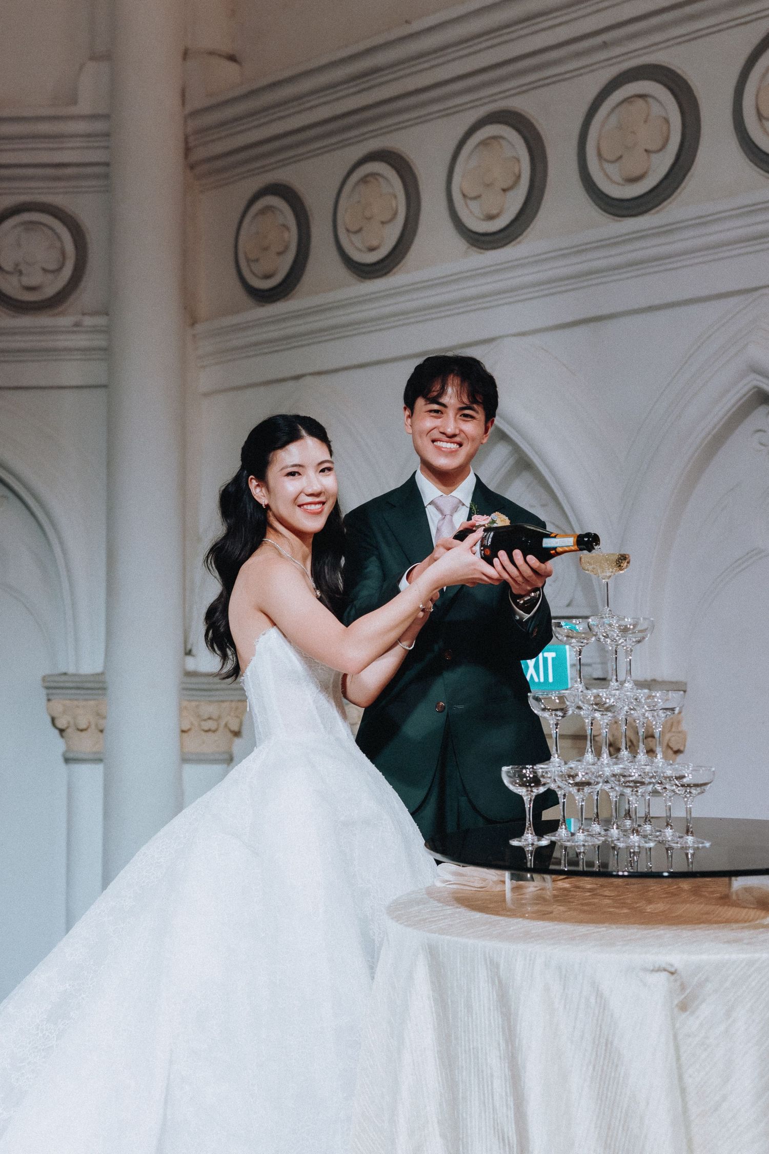 Smiling newlyweds pour champagne into a tower of glasses at their elegant wedding reception.