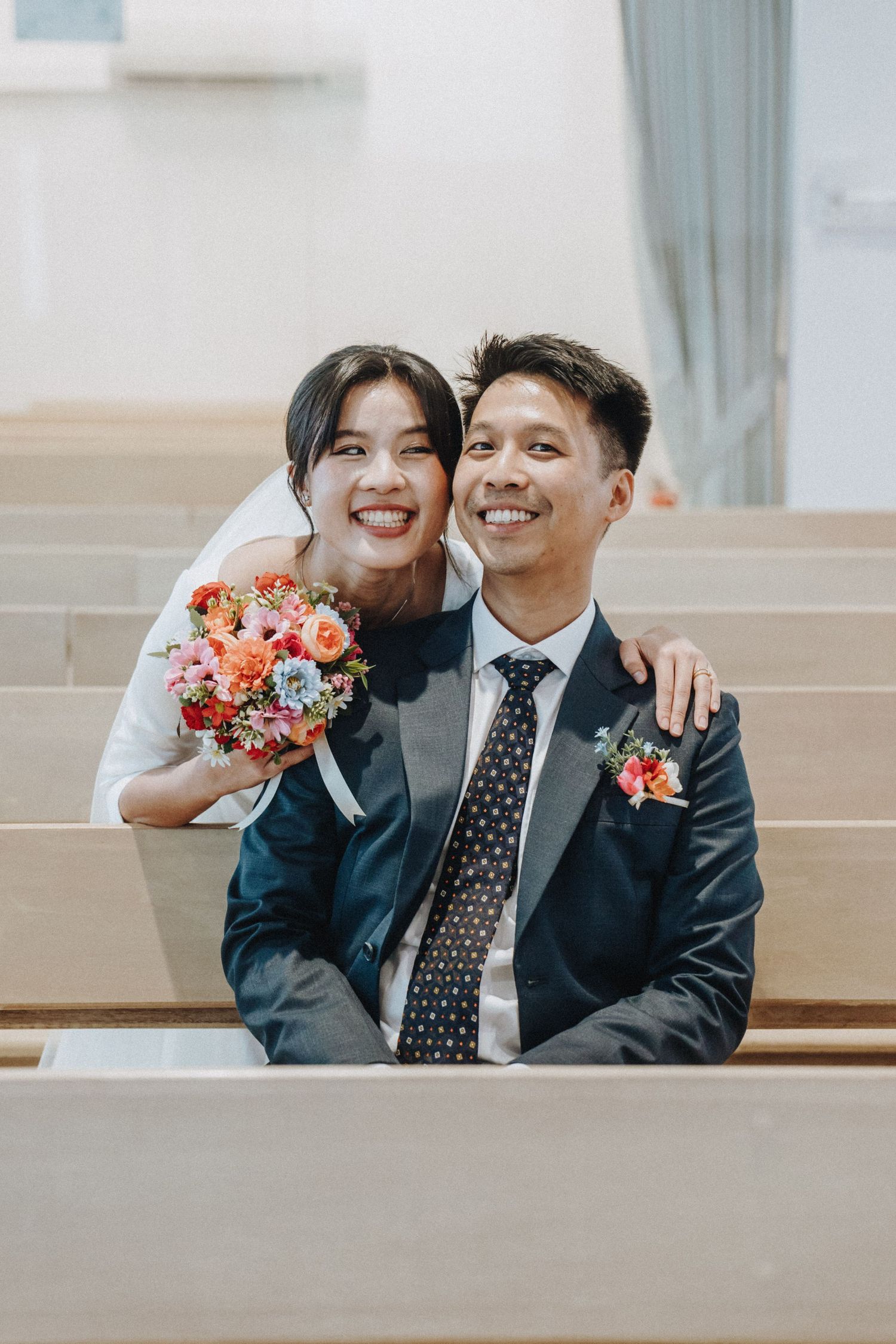 Happy bride and groom smiling together on church pew with colorful bridal bouquet.