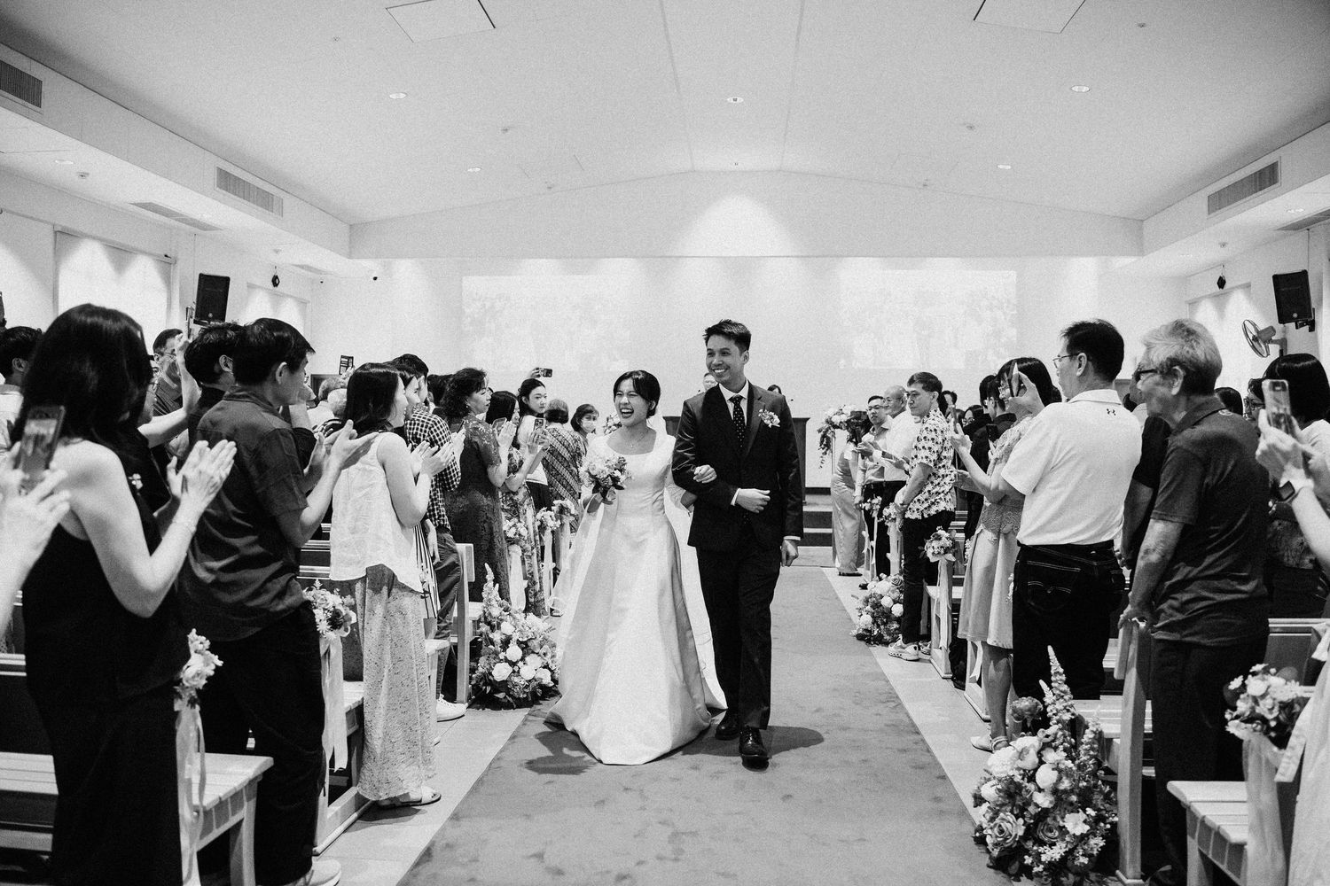 A black and white photo of a bride and groom walking down the aisle as guests clap.