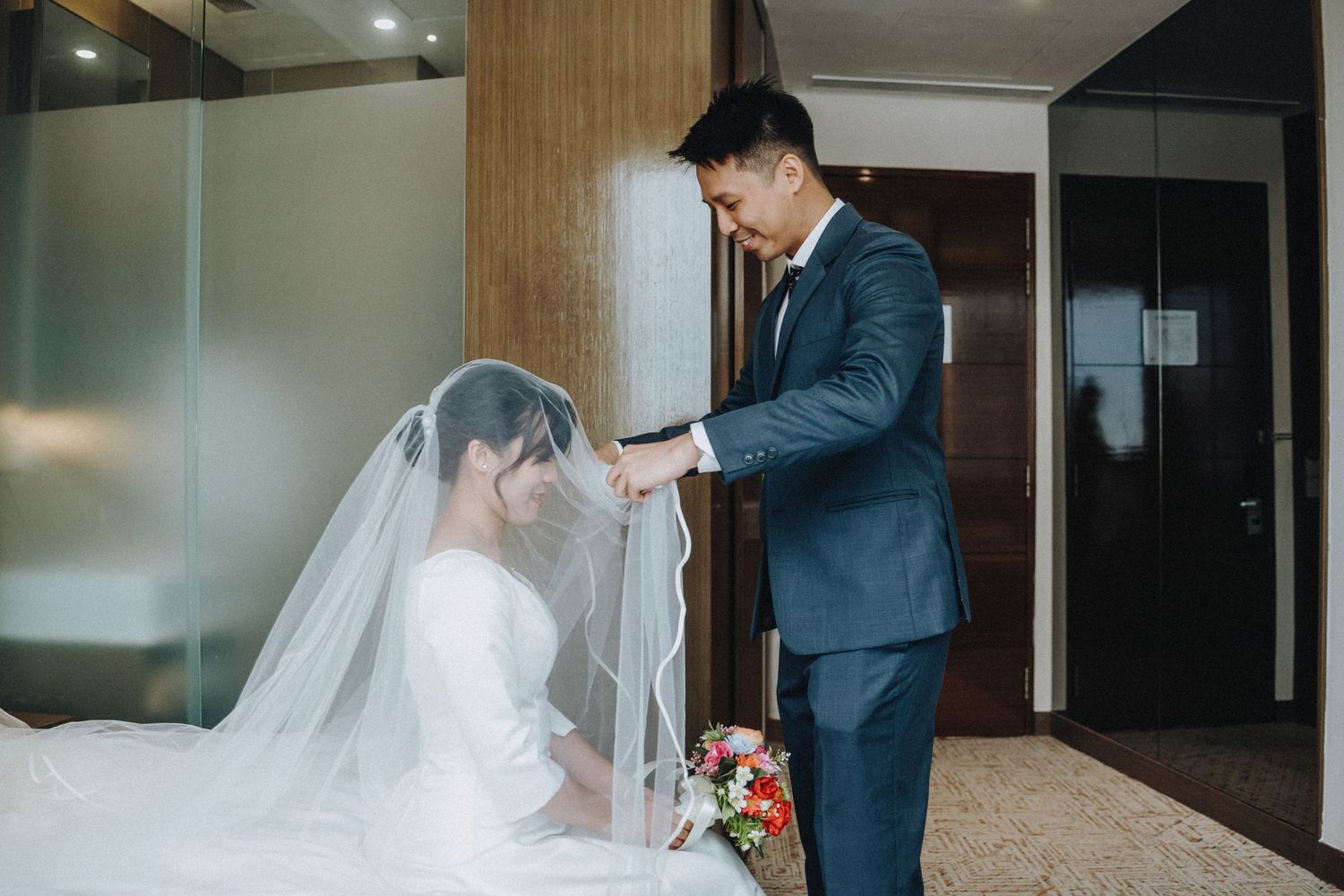 Groom in navy suit adjusts brides veil in hotel room before their wedding ceremony.