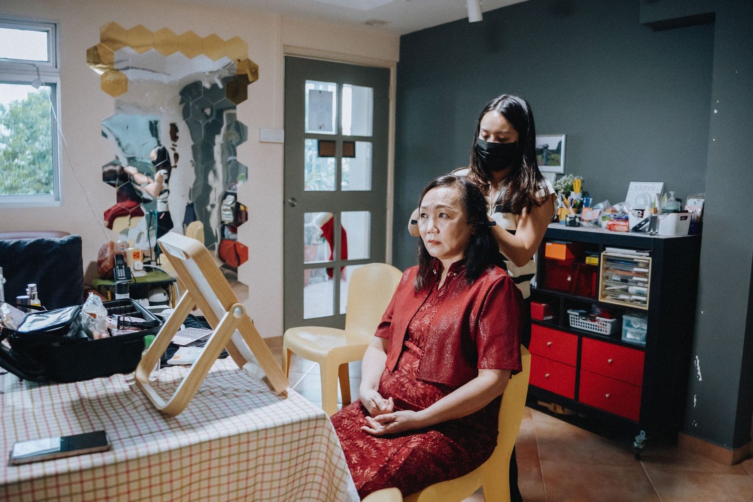 A woman in a red dress sits in a salon chair while a stylist applies makeup in a cozy beauty studio.