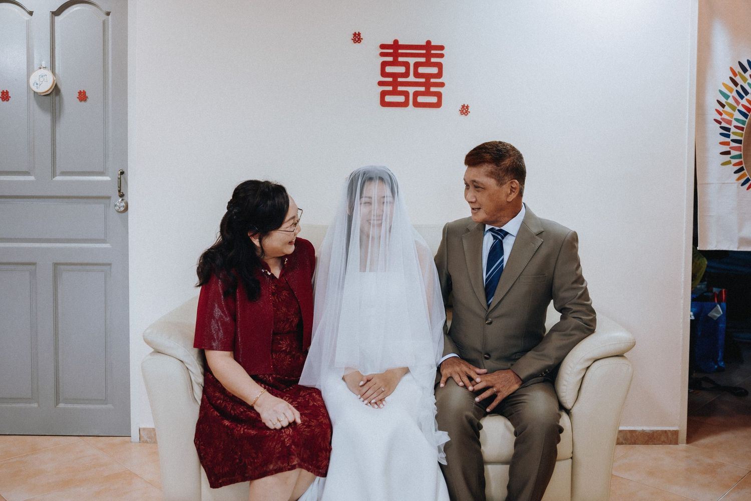 Bride in white veil sits with family during traditional Chinese wedding ceremony, double happiness symbol on wall.