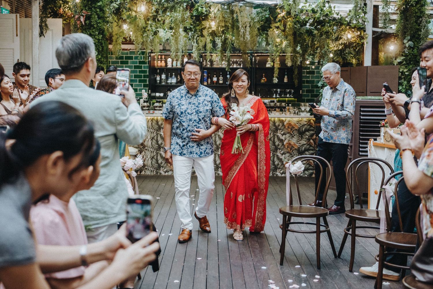 Couple in traditional Indian and Hawaiian attire walk down the aisle at an outdoor wedding ceremony.