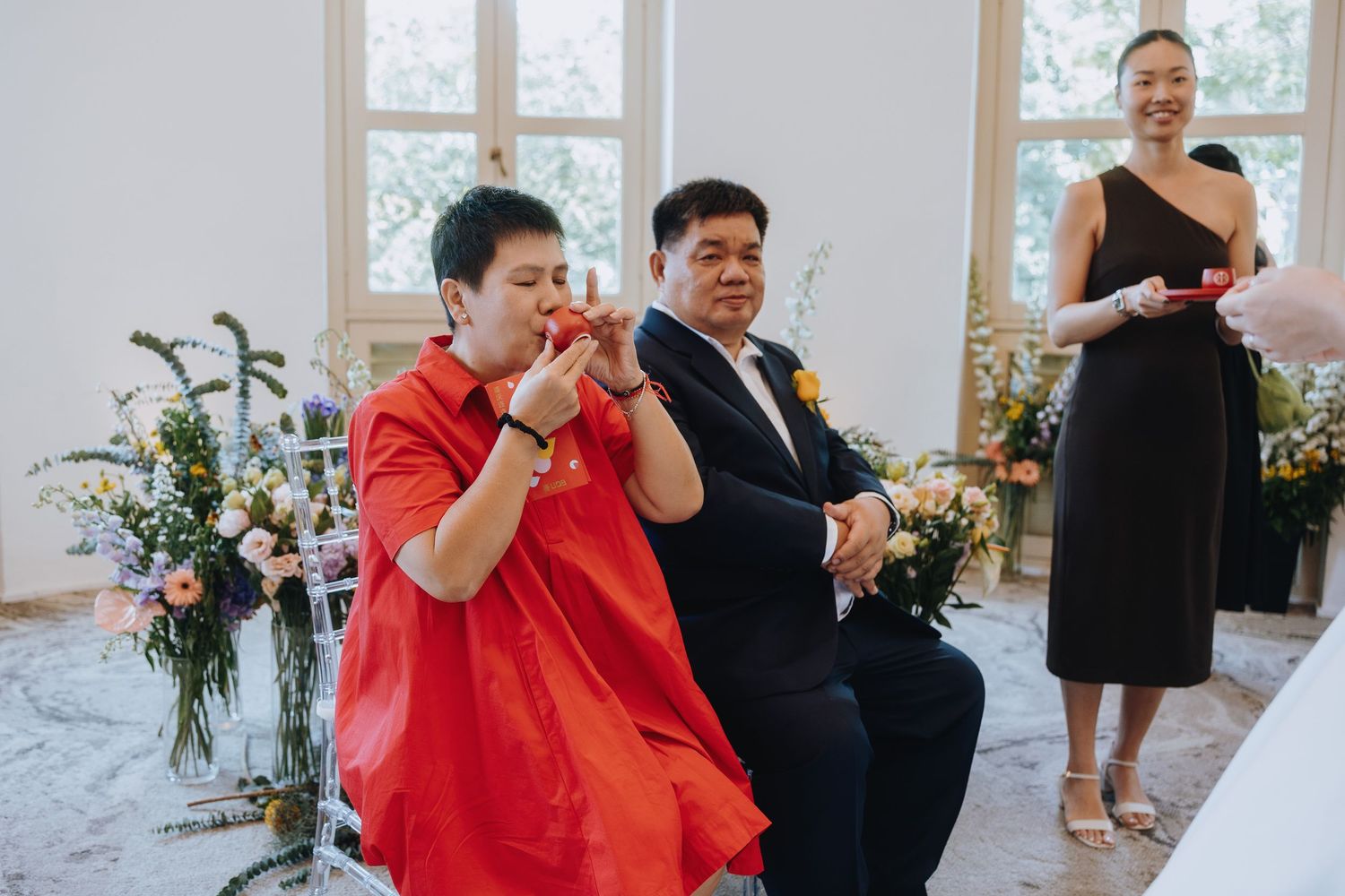 Woman in red dress and man in yellow tie participate in a traditional wedding tea ceremony indoors.