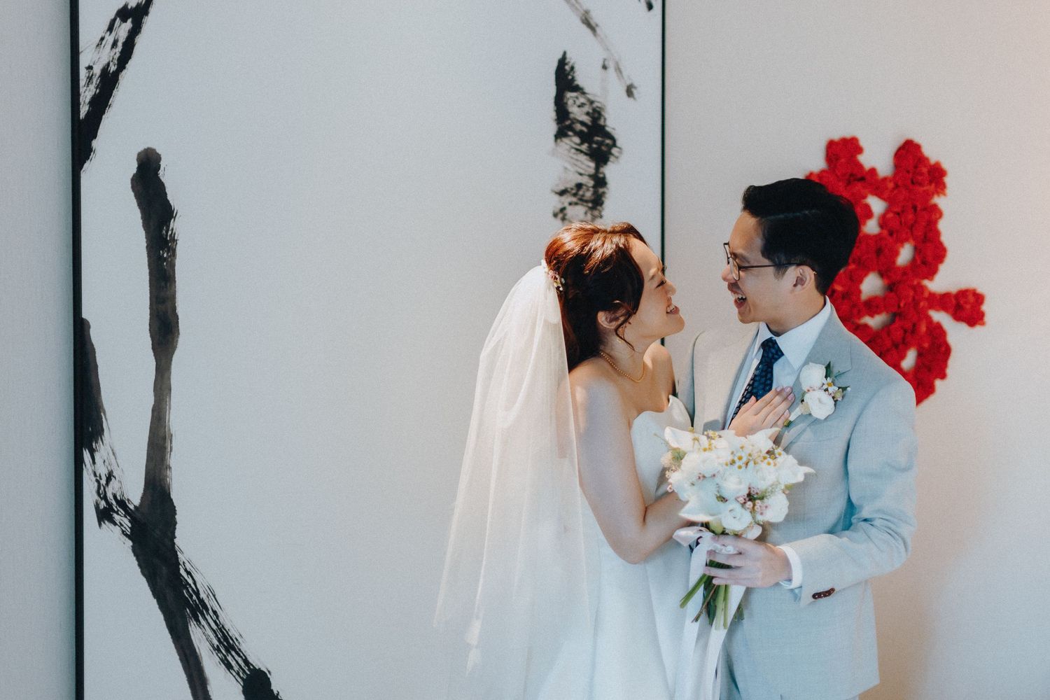 Wedding couple shares candid moments against white wall with vibrant red floral accents.