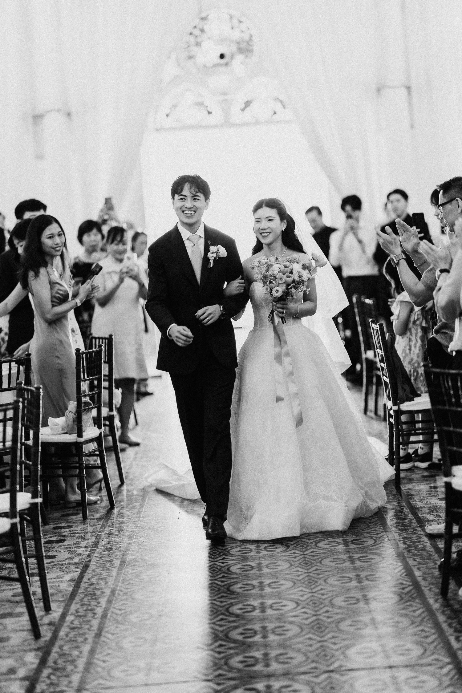 Black and white photo of smiling newlyweds walking down the aisle at their wedding ceremony.
