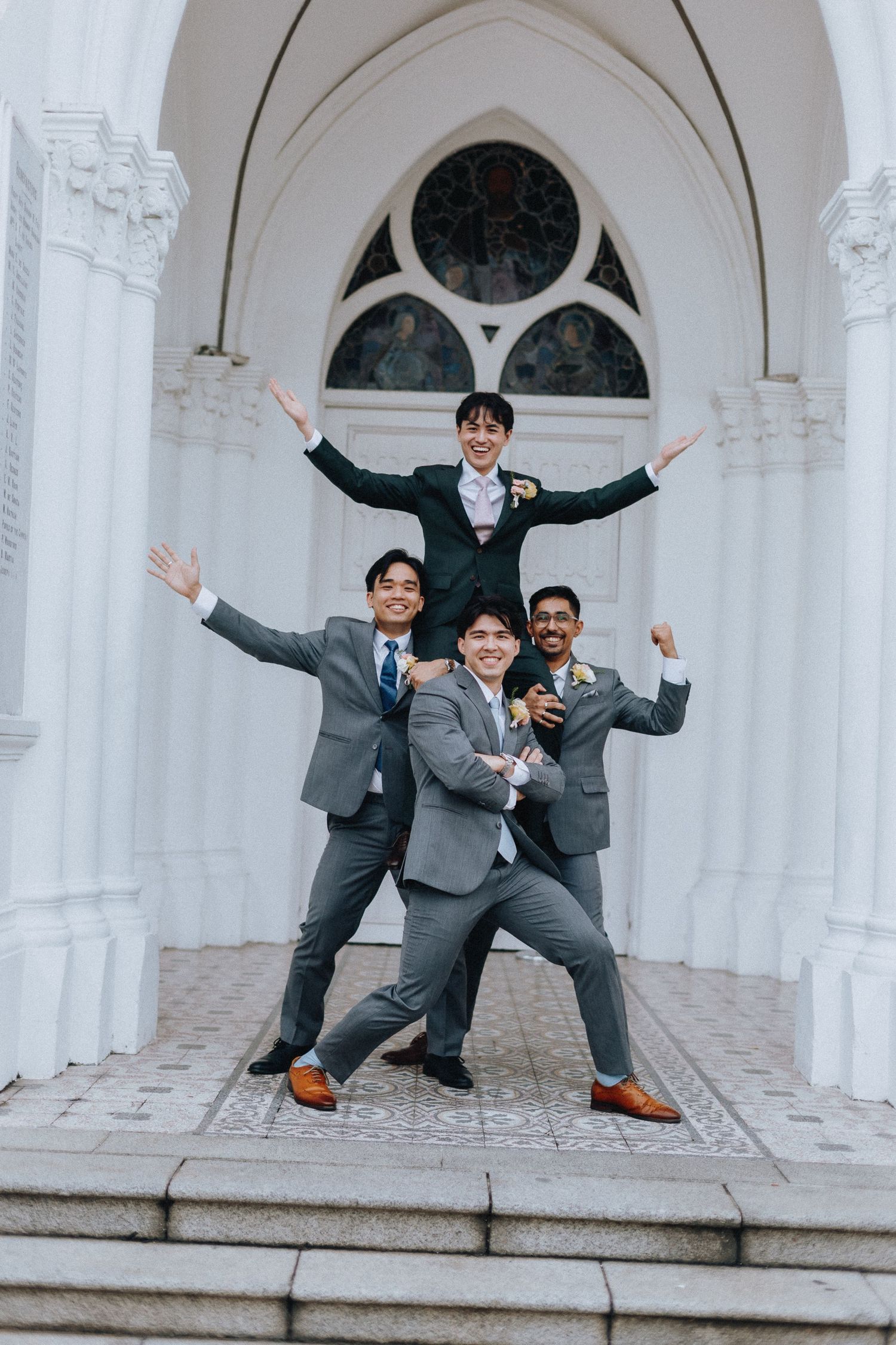 Four groomsmen in suits posing playfully on church steps, one lifted on shoulders with arms raised in celebration.