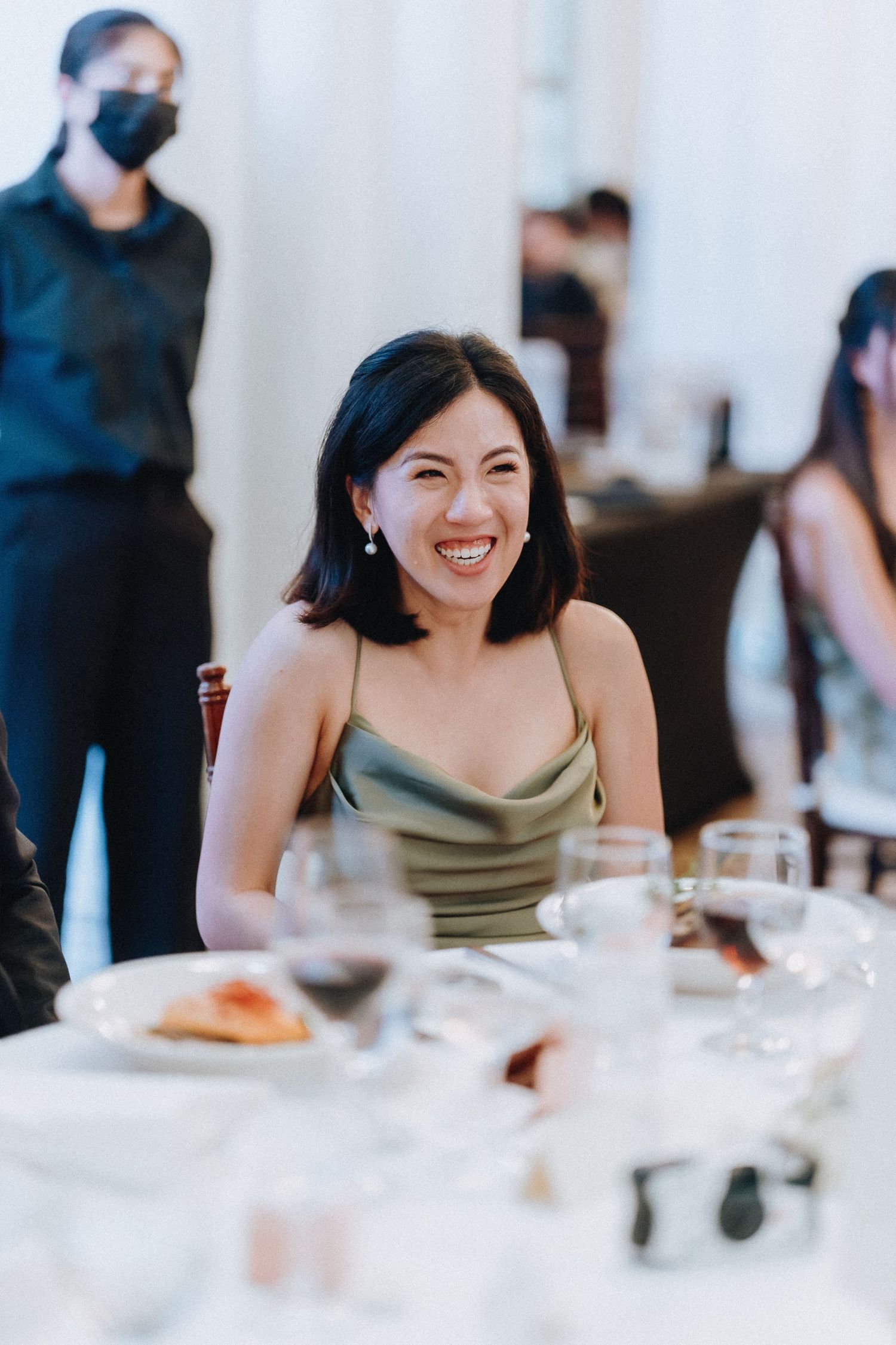 Smiling woman in elegant olive dress seated at a wedding reception dinner table.