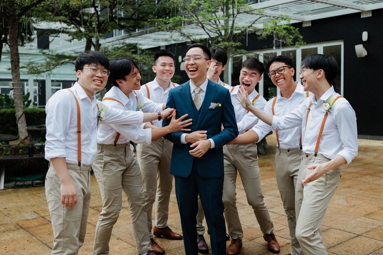 Laughing groom in navy suit surrounded by cheerful groomsmen in khaki pants with orange suspenders at outdoor wedding celebration.