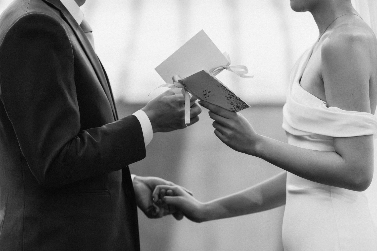 Black and white close-up of bride and groom exchanging vow booklets and holding hands during wedding ceremony.