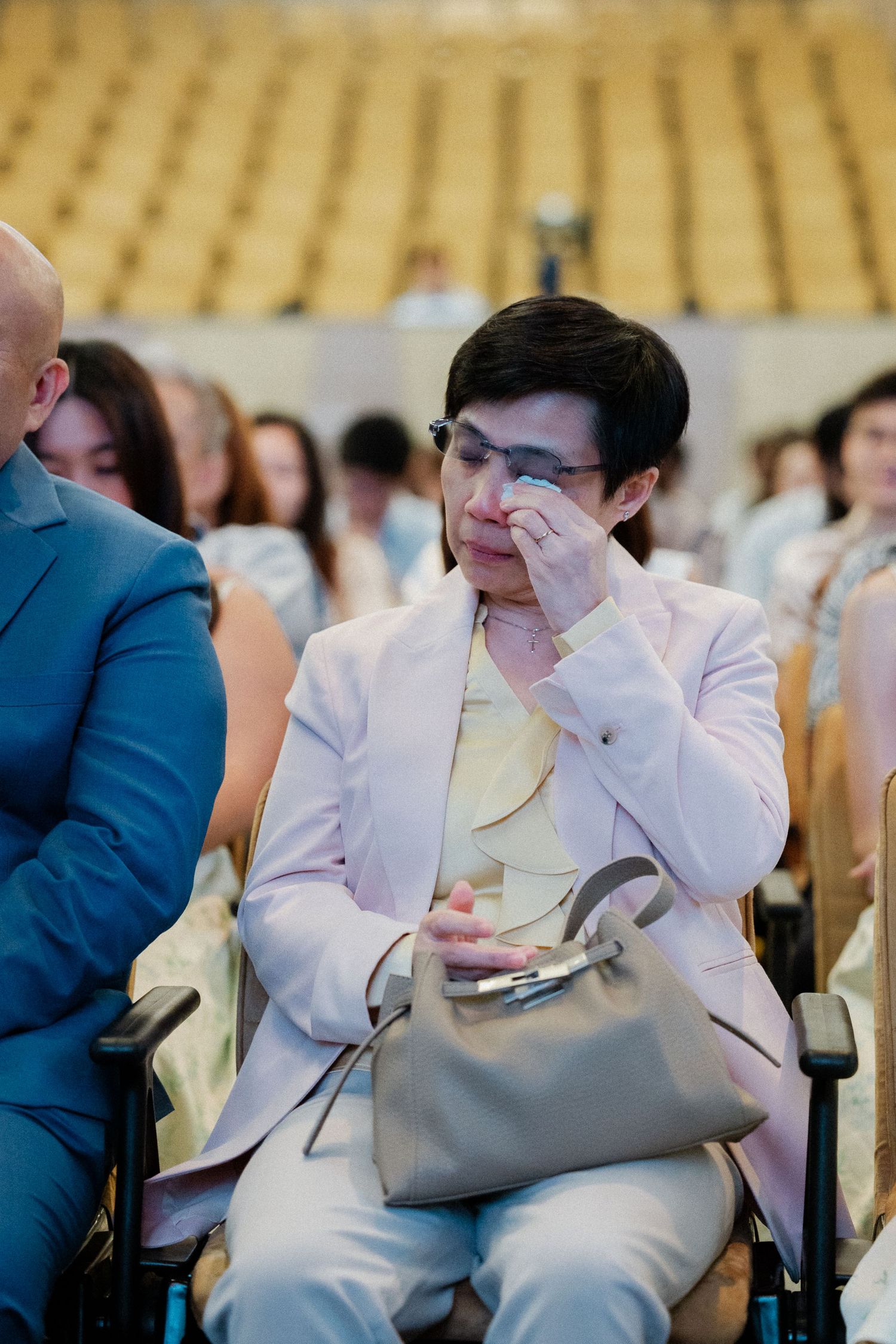 Emotional mother in pink blazer wiping tears during wedding ceremony seated in auditorium.