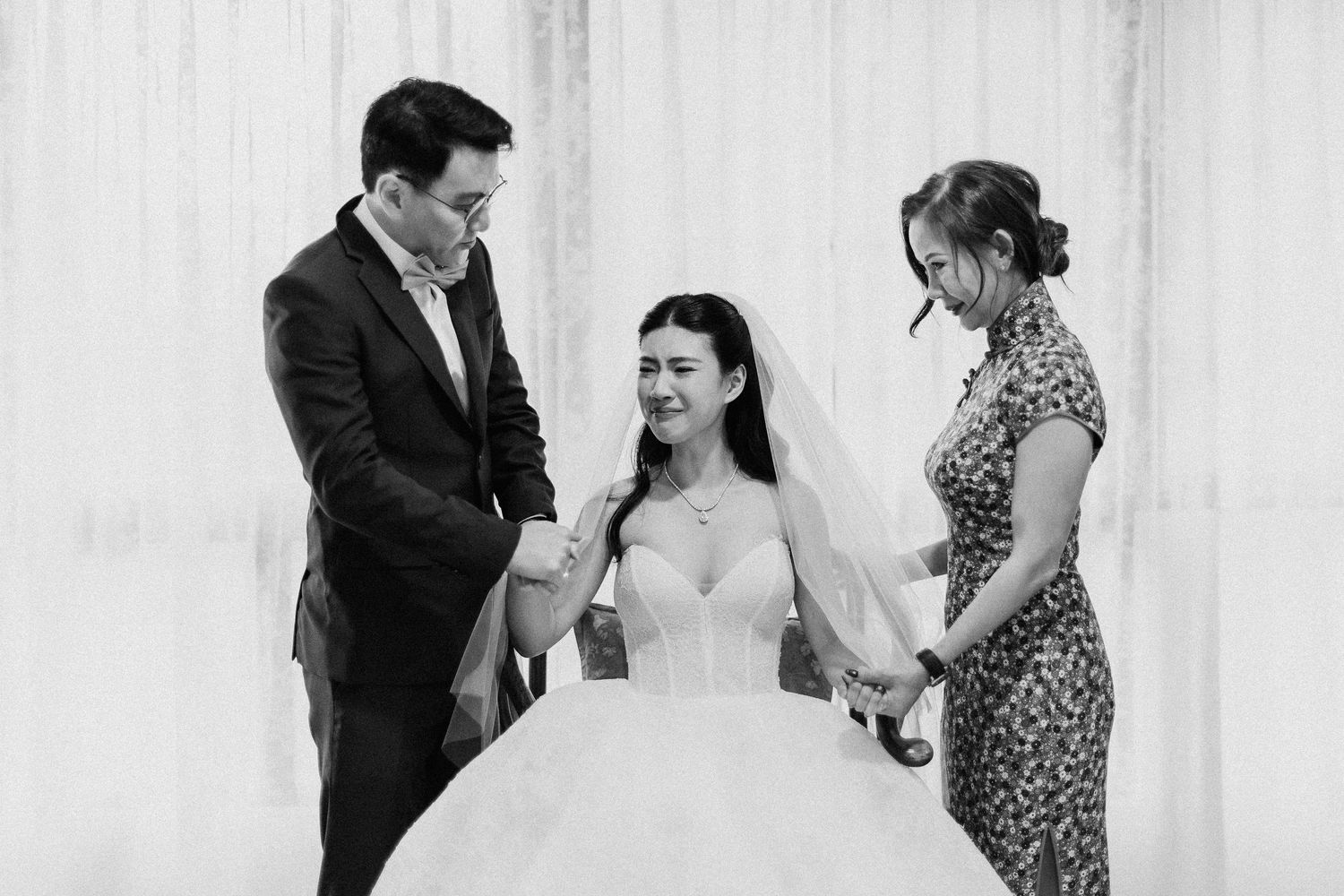 Bride in white wedding gown and veil smiling with groom and female assistant in black and white photo.