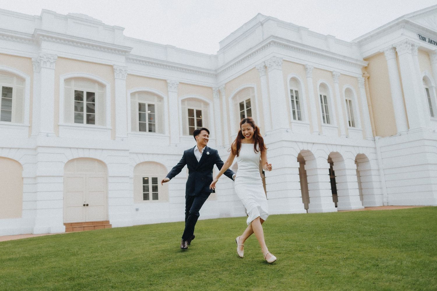 Couple runs playfully across a lawn in front of a grand white colonial building.