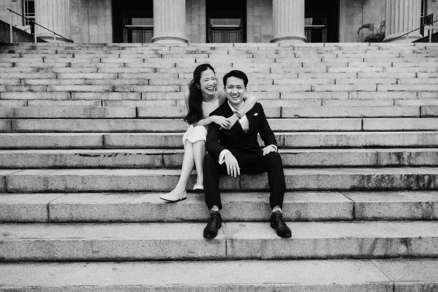 A couple sits together on stone steps of a grand building with columns in black and white.