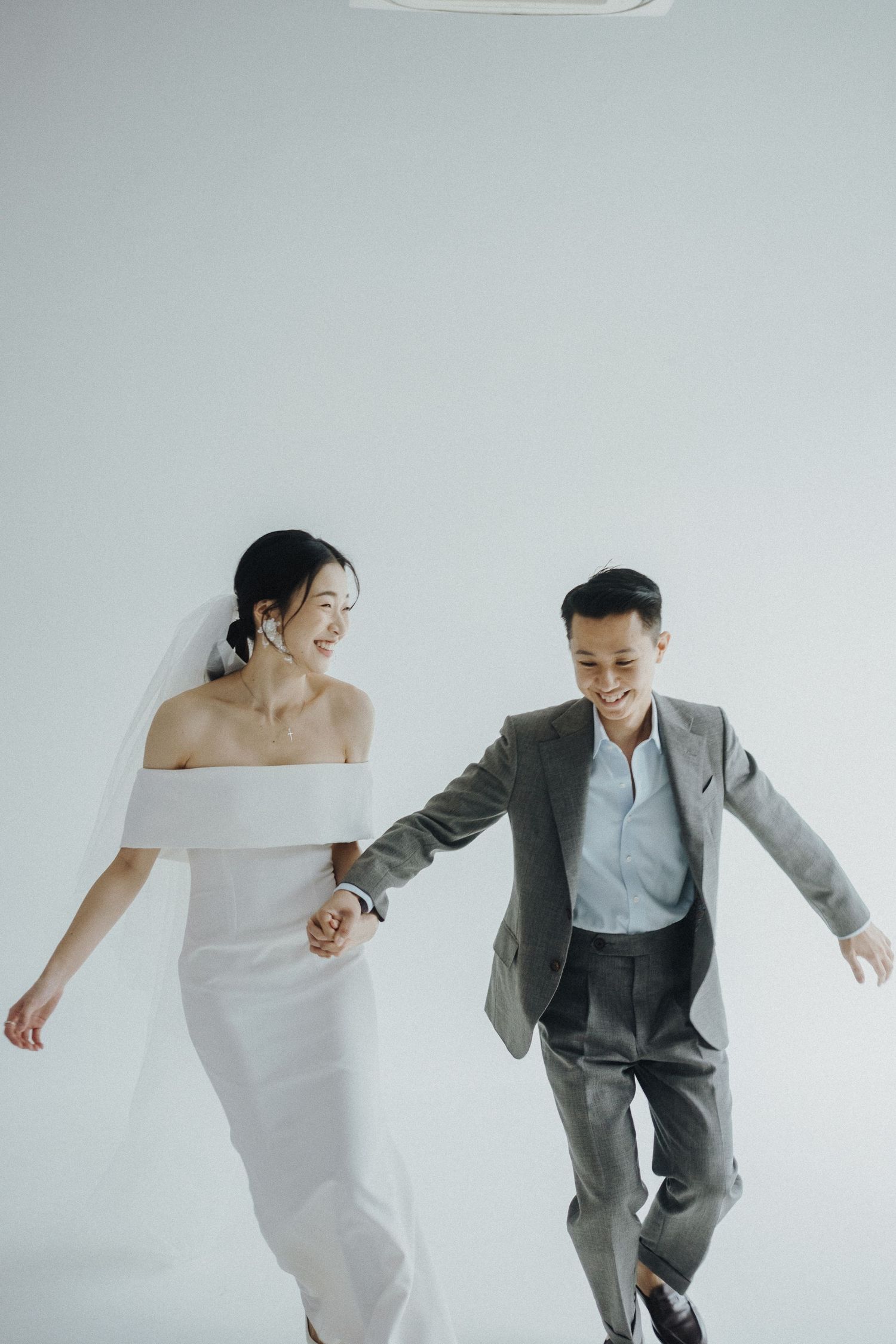 A couple in wedding attire holding hands and walking joyfully together against a minimalist white background.