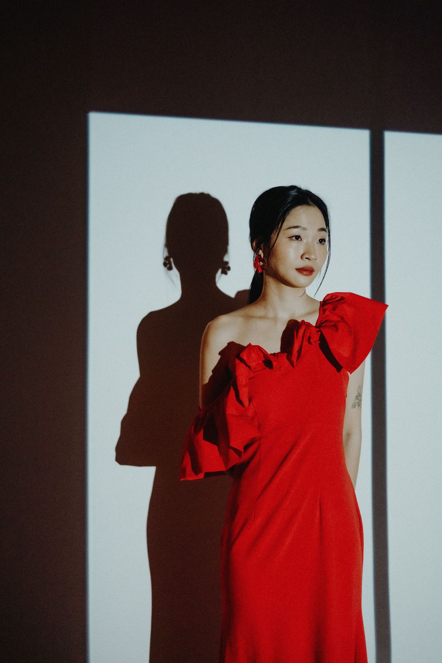 Woman in vibrant red one-shoulder dress poses against white wall creating dramatic shadow silhouette.