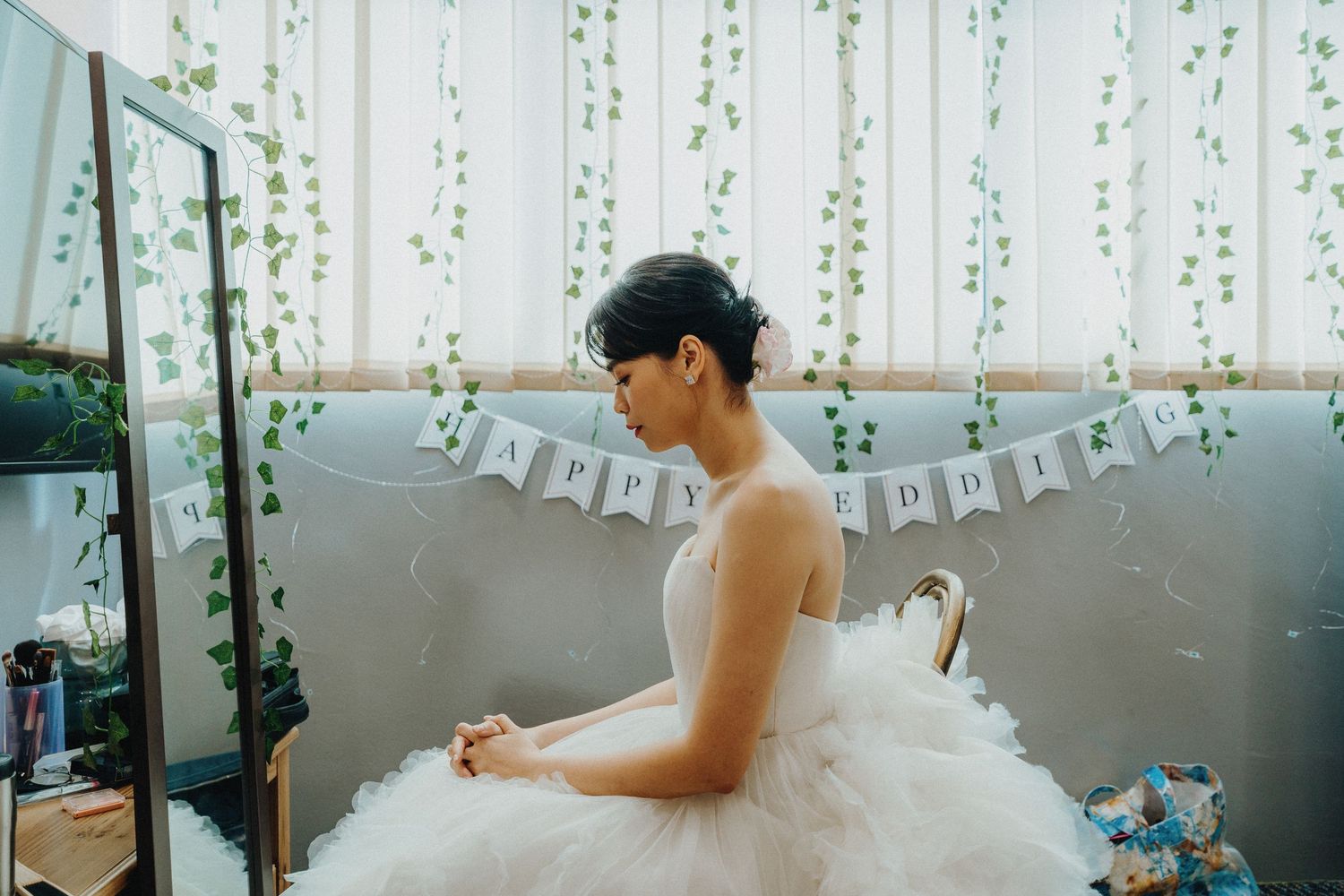 A person in a white tulle wedding dress adjusts details while sitting on a gray couch with banners.