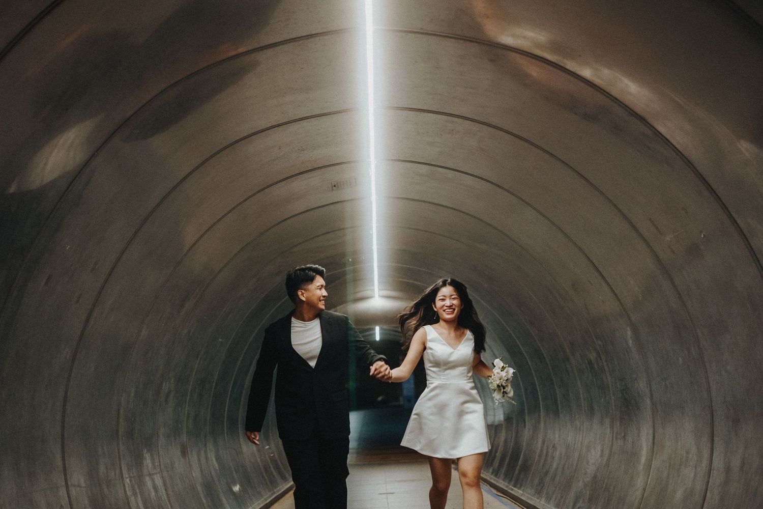 A wedding couple runs playfully through a circular concrete tunnel illuminated by a bright light beam.