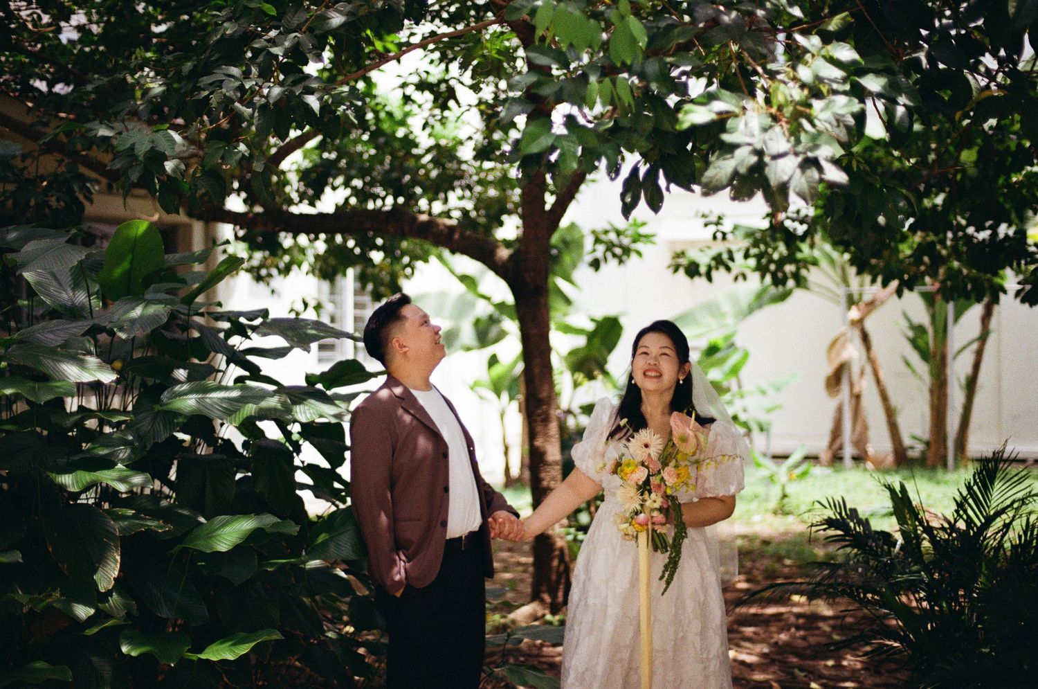 Wedding ceremony guests gather in a shaded garden setting with lush greenery and natural decor.