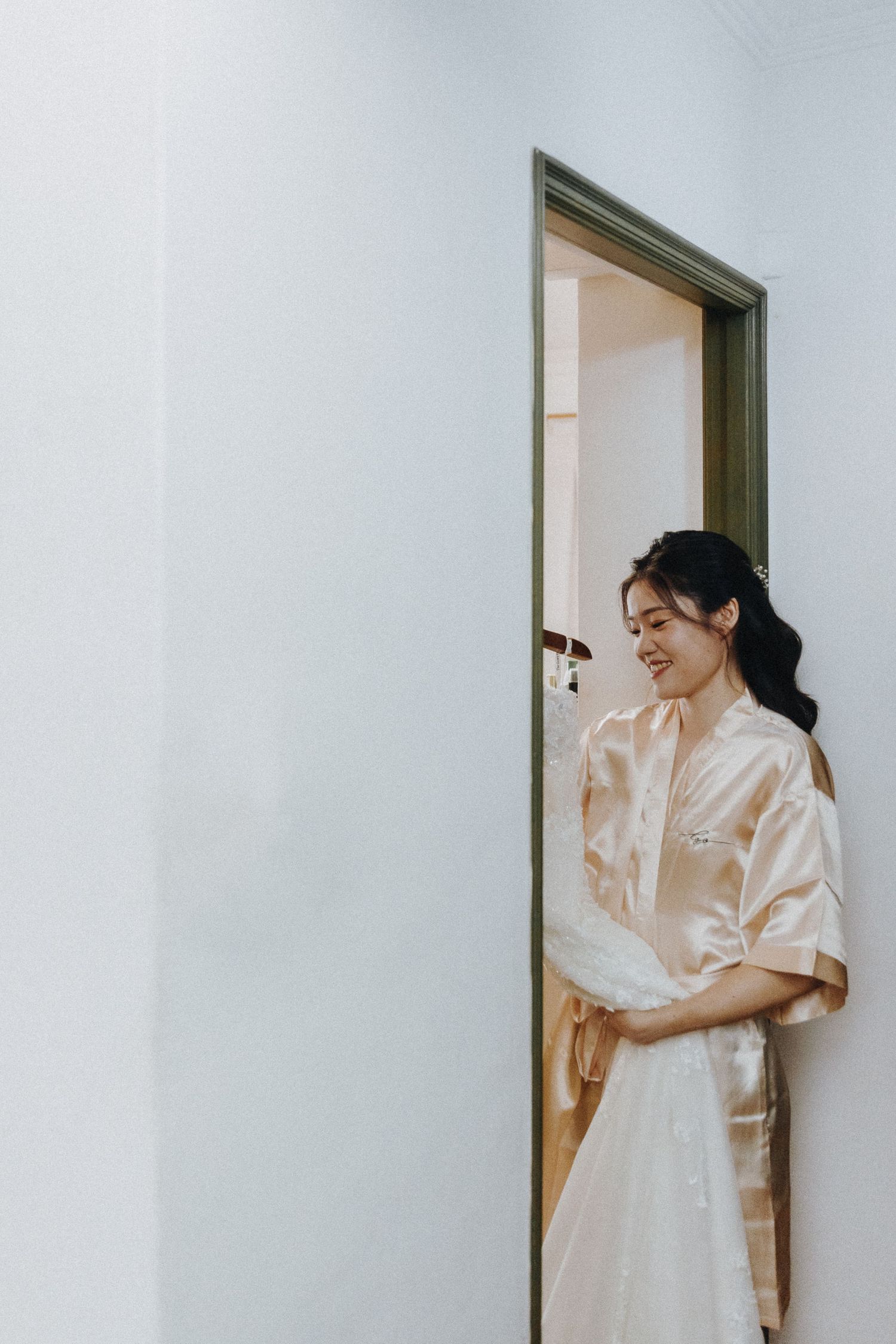 A bride in a silk robe stands thoughtfully against a white wall and doorway during wedding preparations.
