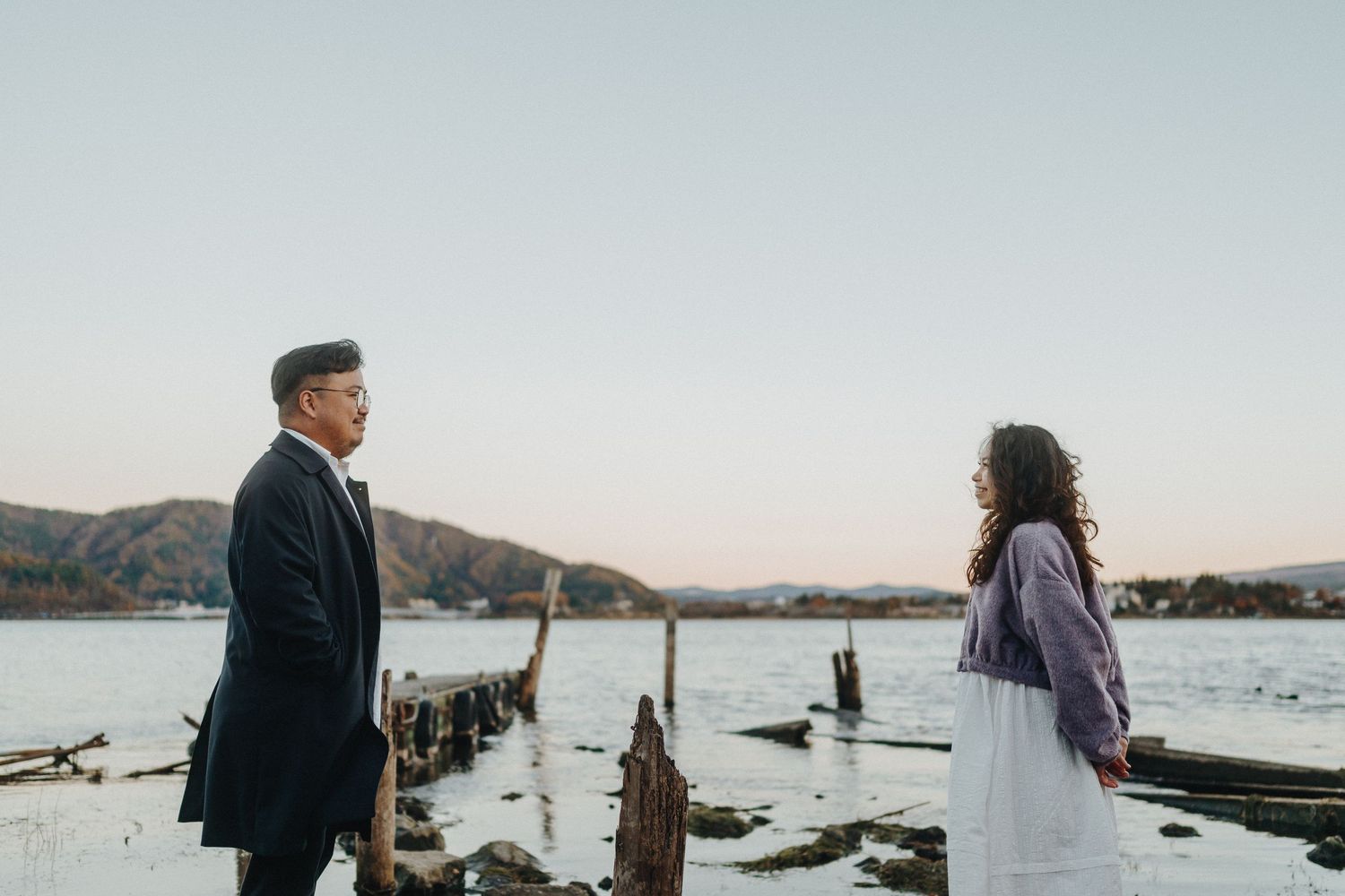 Couple in formal attire standing by waterfront with mountains at sunset.