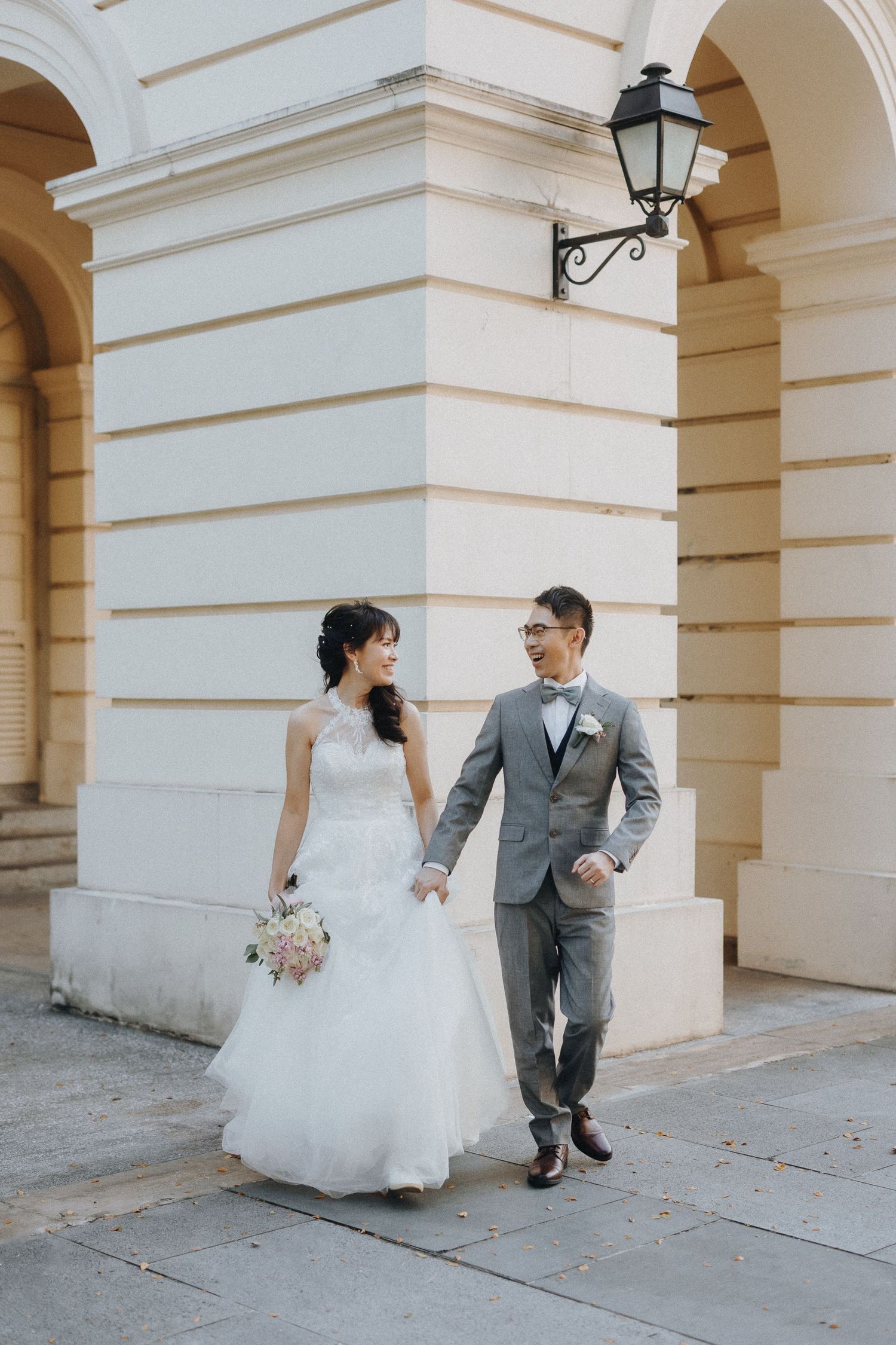 Elegant couple in wedding attire walking through ornate arched colonnade, creating romantic candid moments.
