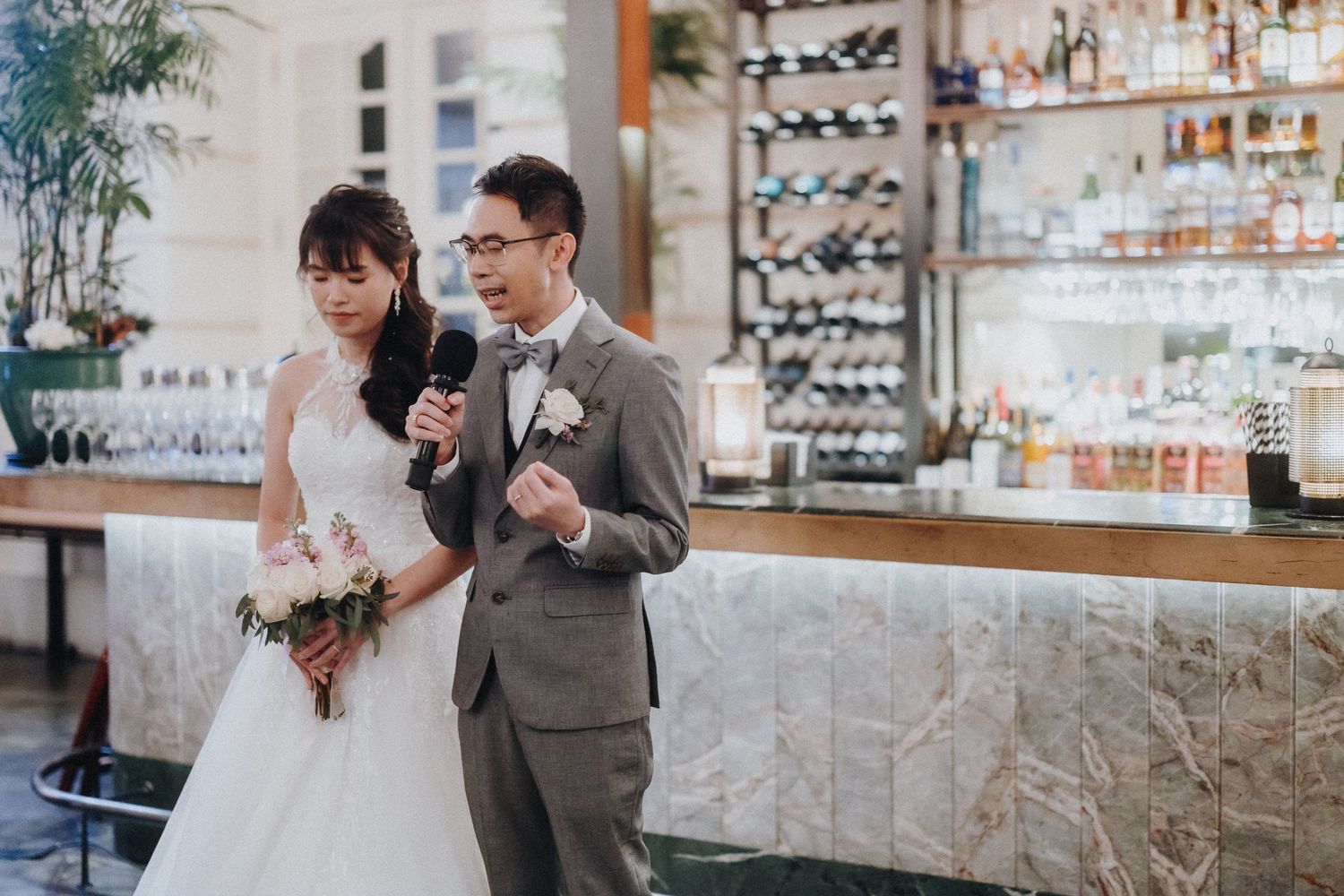 Newlywed couple stands together at an elegant bar with wine bottles on display in the background.