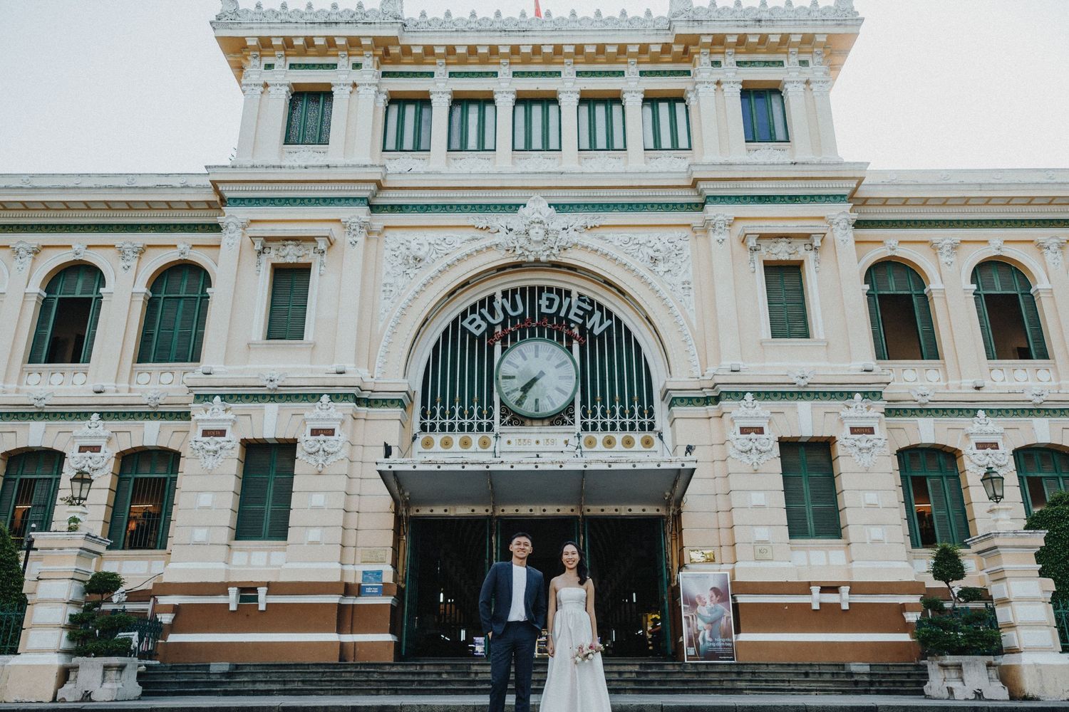 A couple stands in front of the ornate colonial-style Saigon Central Post Office with its grand arched entrance and clock.