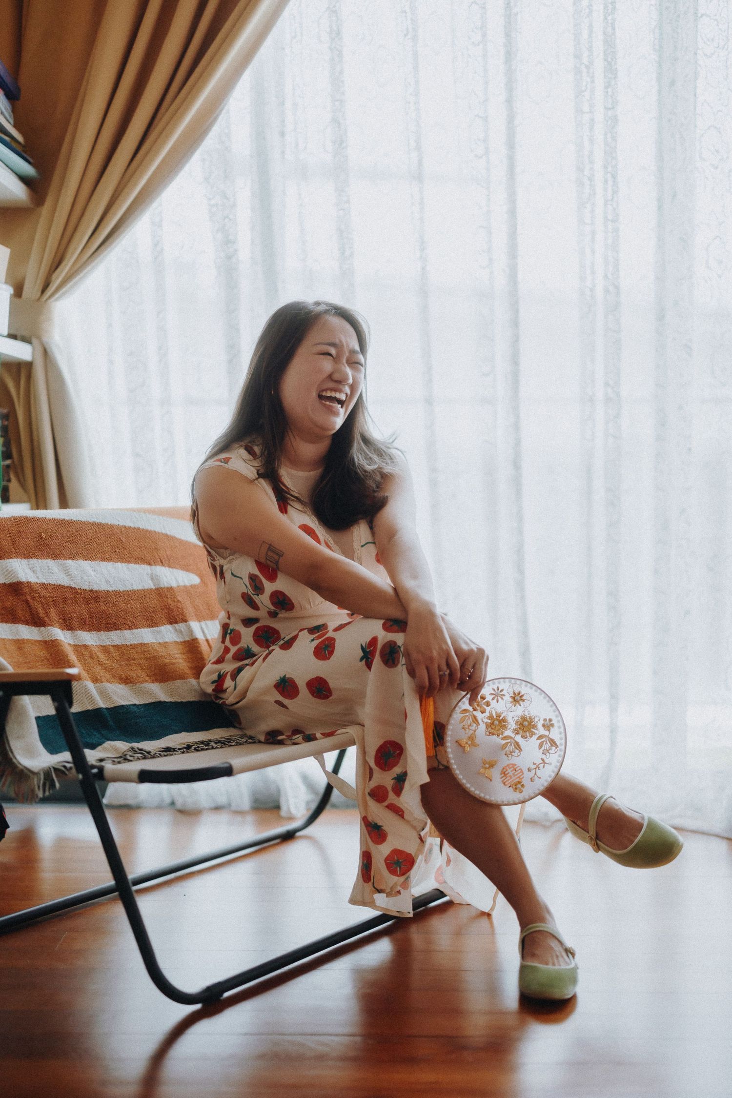 A person in a polka dot dress laughs while sitting on a folding chair indoors by a window.