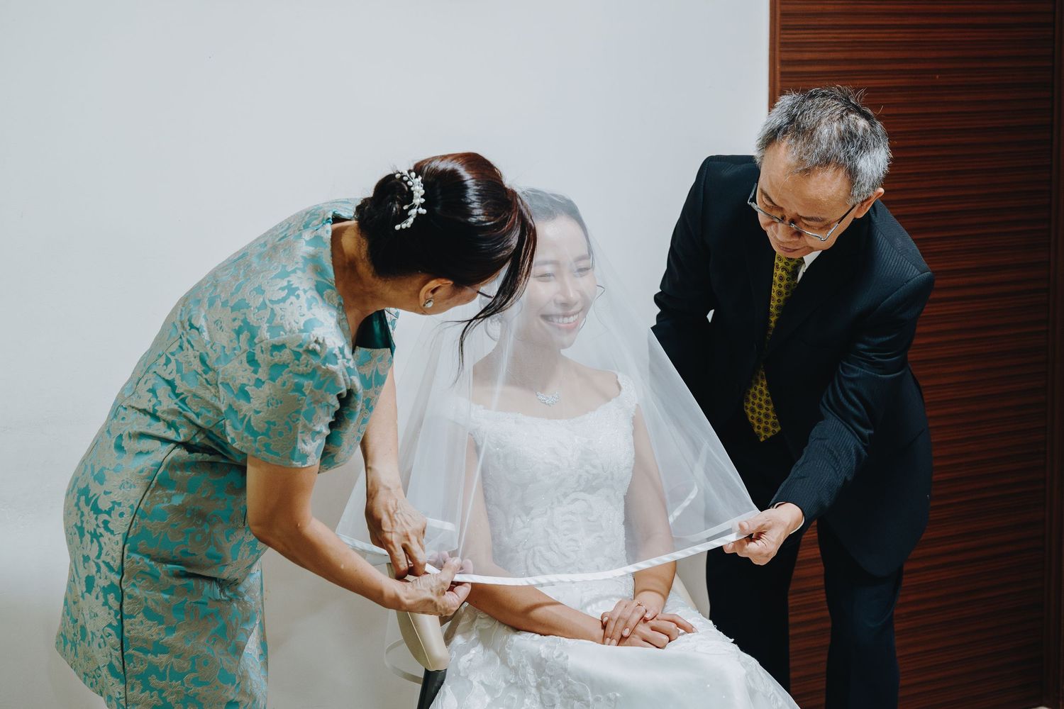 A bride in white wedding dress and veil being helped with final preparations by two attendants in a bright indoor setting.