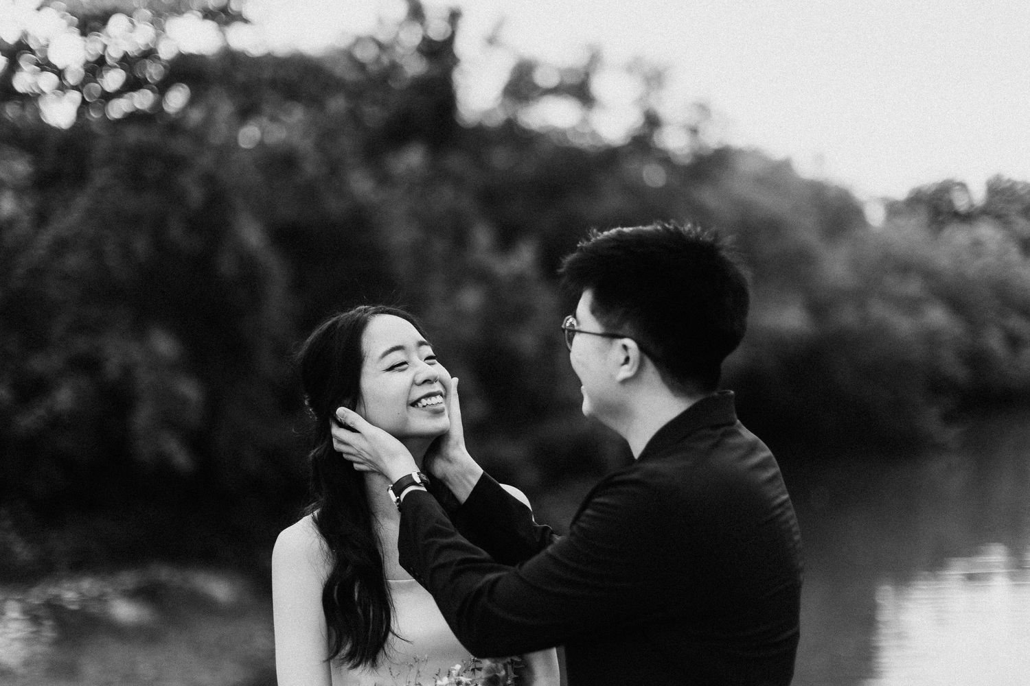 Two people share a tender moment while laughing by a lake in black and white.
