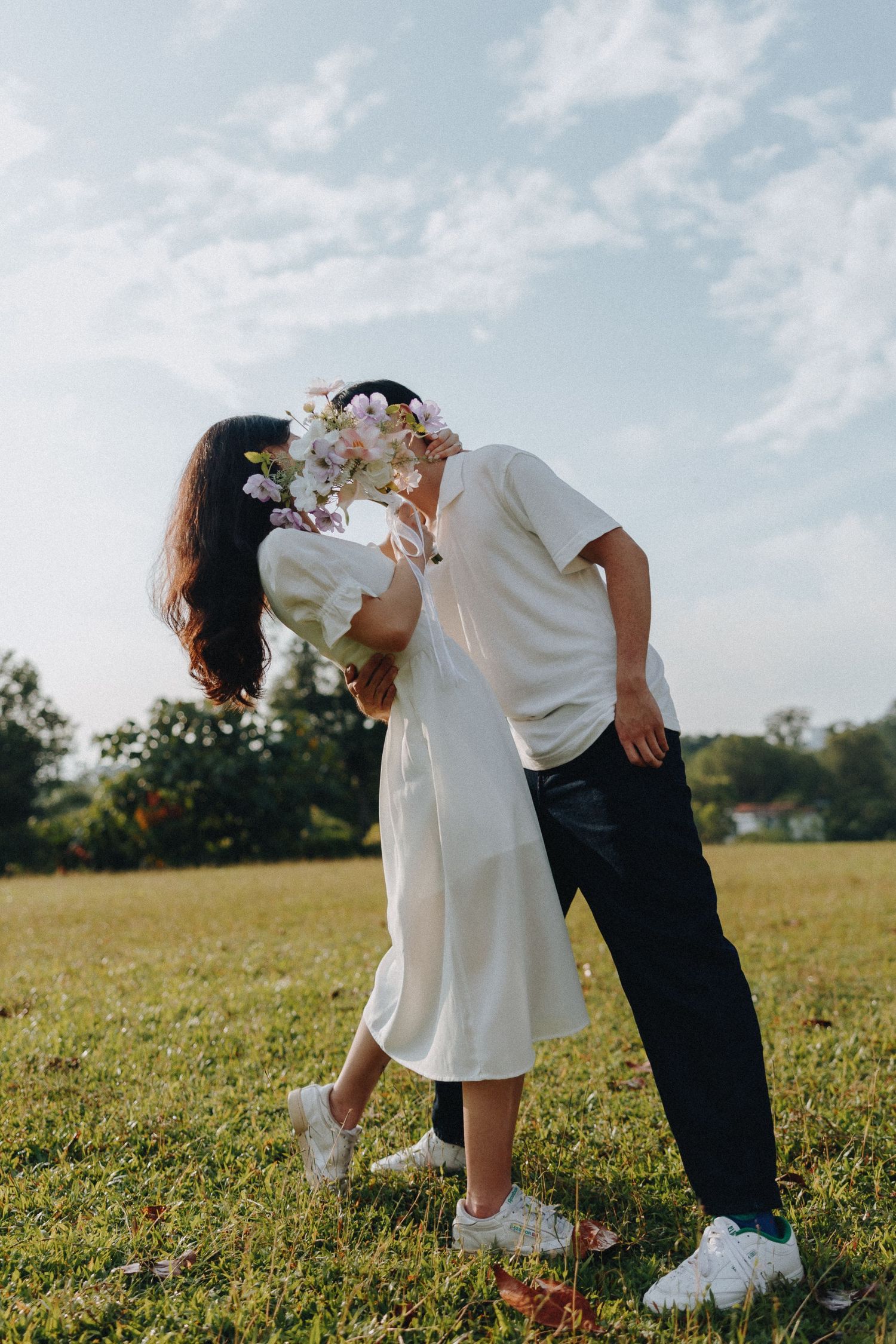 A couple in white shares a romantic moment with flowers in an outdoor setting.