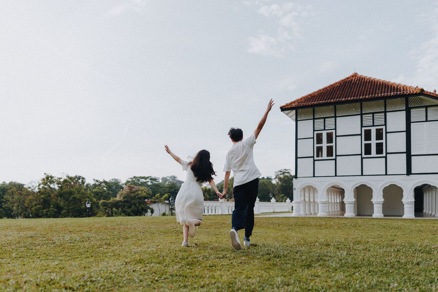 A couple runs joyfully across a lawn towards a colonial-style building.