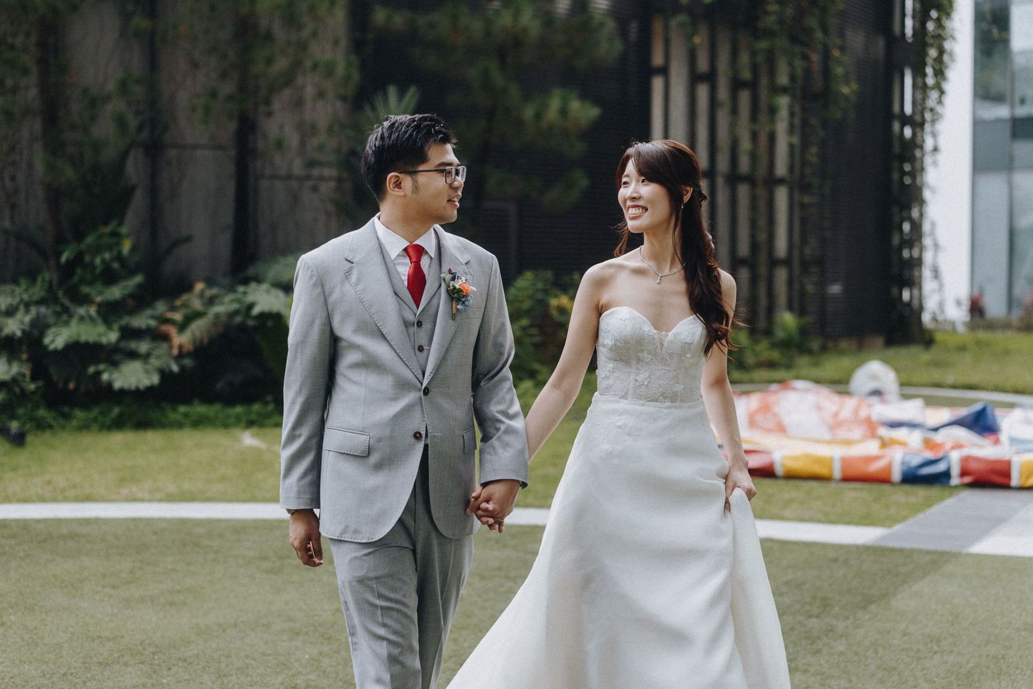 Happy newlywed couple holds hands while walking together on grass, bride in white gown and groom in light grey suit.