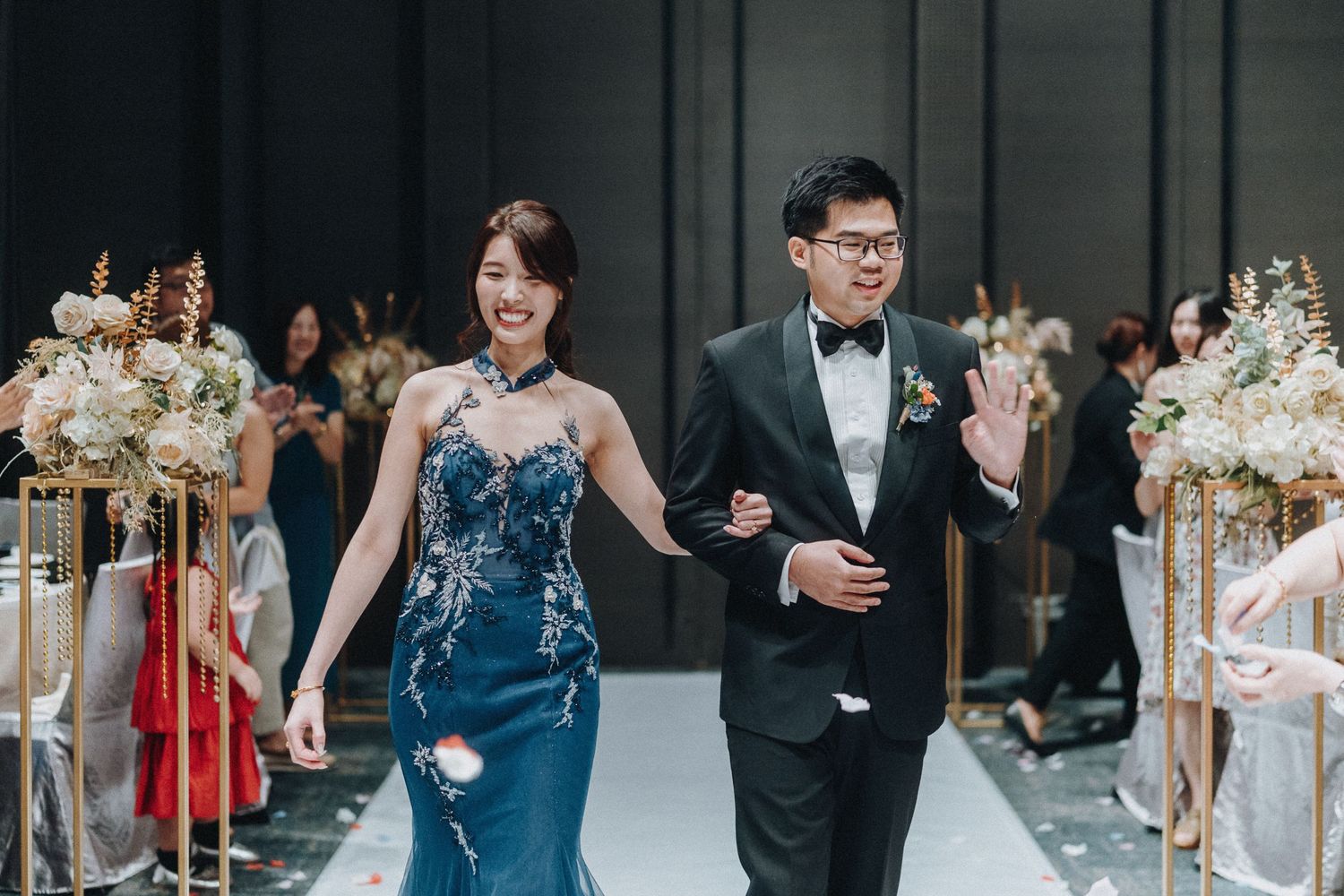 Elegantly dressed couple walk down a modern aisle decorated with floral arrangements at an indoor wedding ceremony.