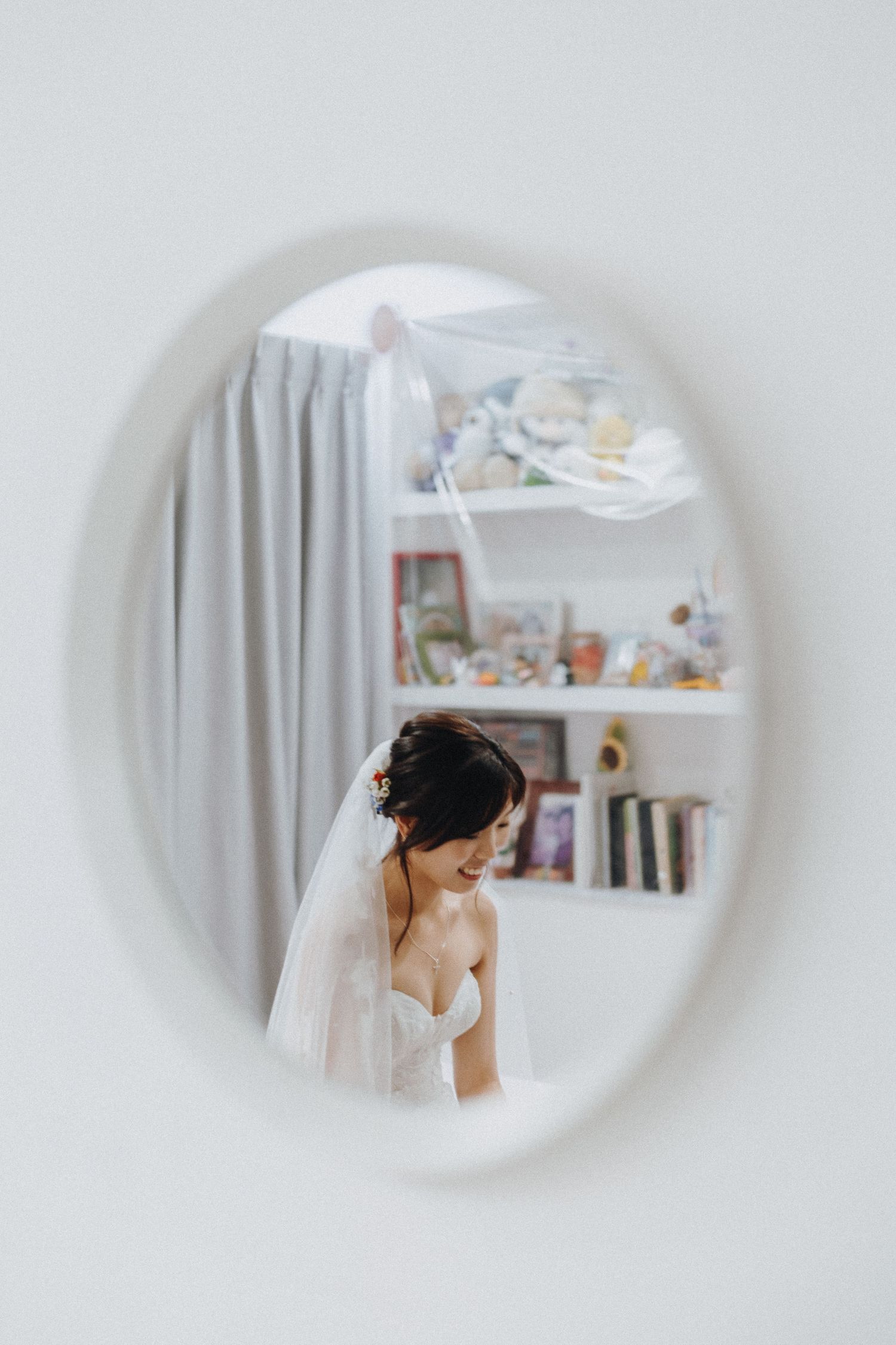 A bride's reflection in an oval mirror shows her wearing a white wedding dress and veil with bookshelves in the background.