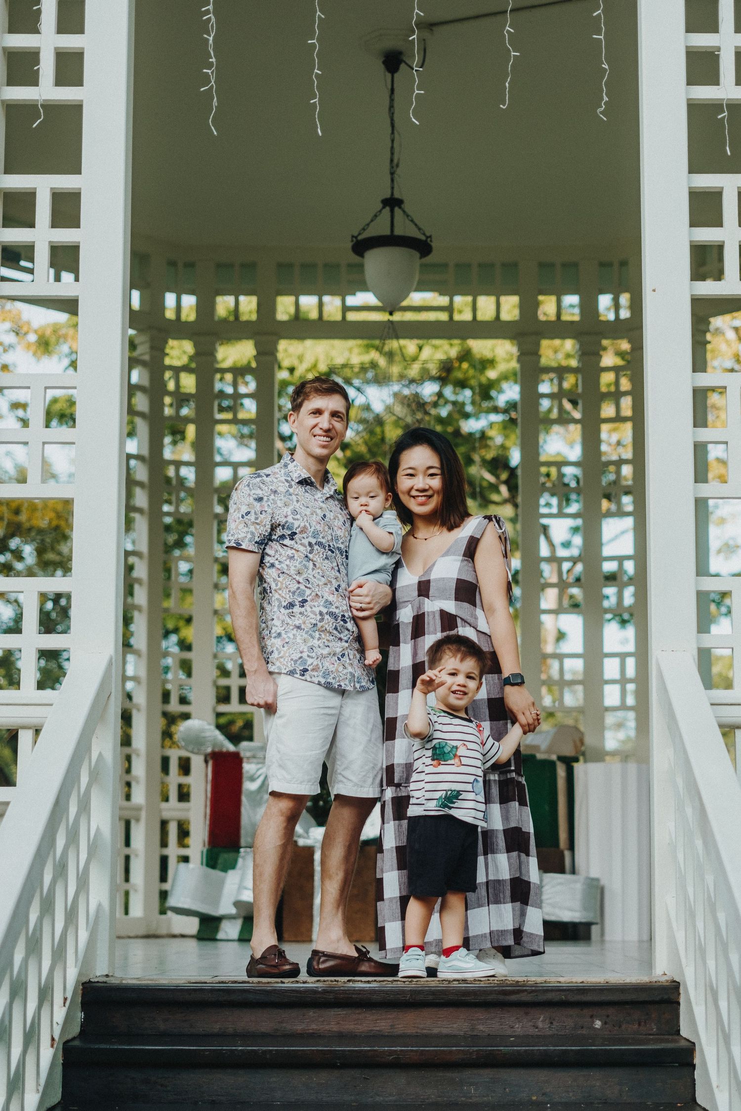 A happy family wearing coordinated outfits pose together on the steps of a white gazebo on a sunny day.