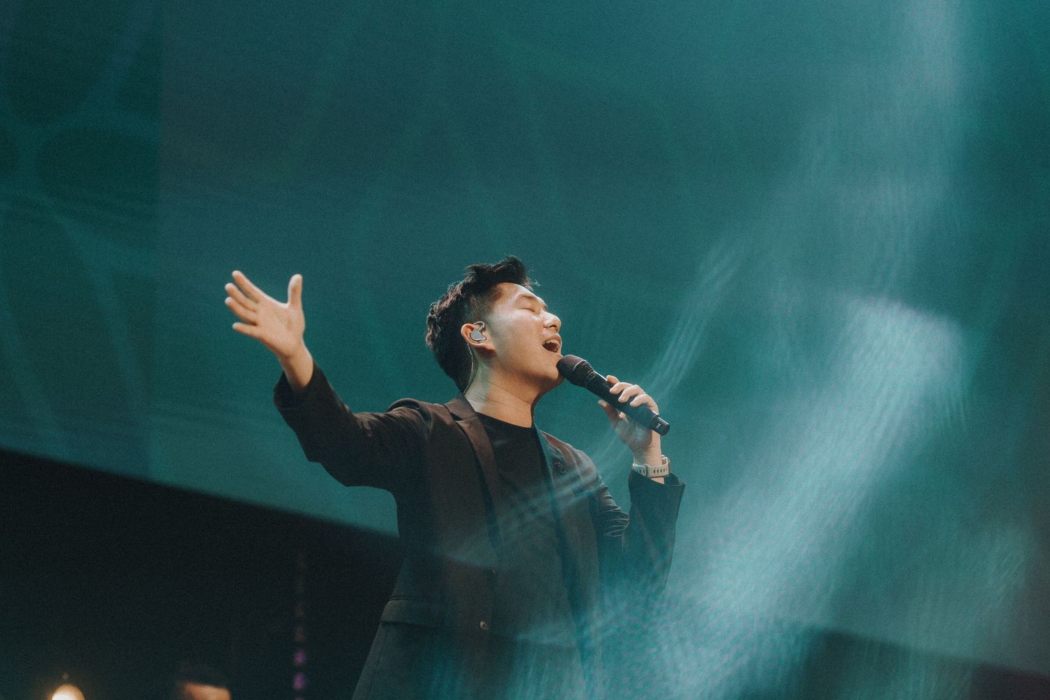 Stage performer raising hands during emotional musical performance with teal lighting and dramatic atmosphere.